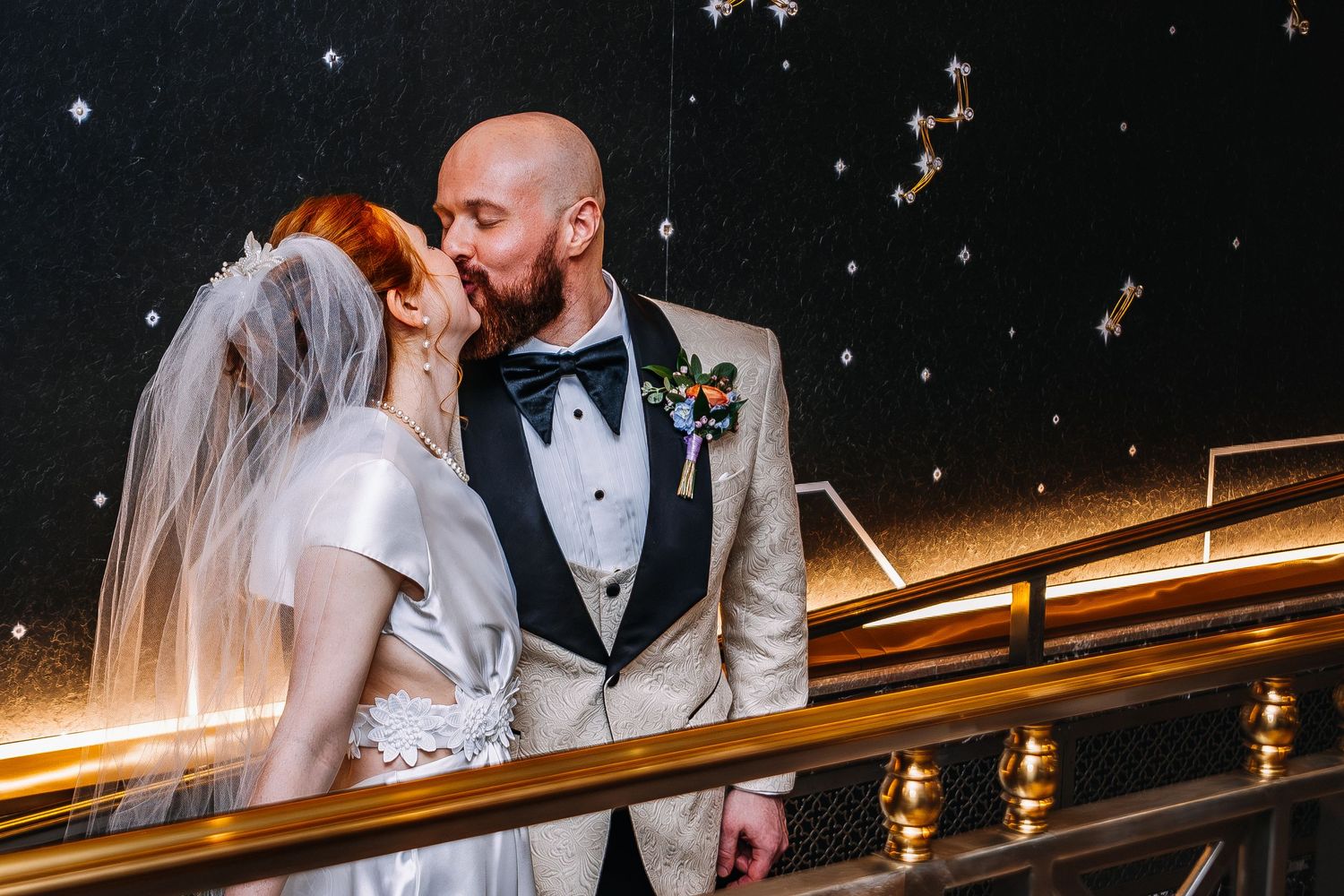 Bride and groom share a romantic kiss on a staircase, groom in gold tuxedo with boutonniere, bride in white veil.