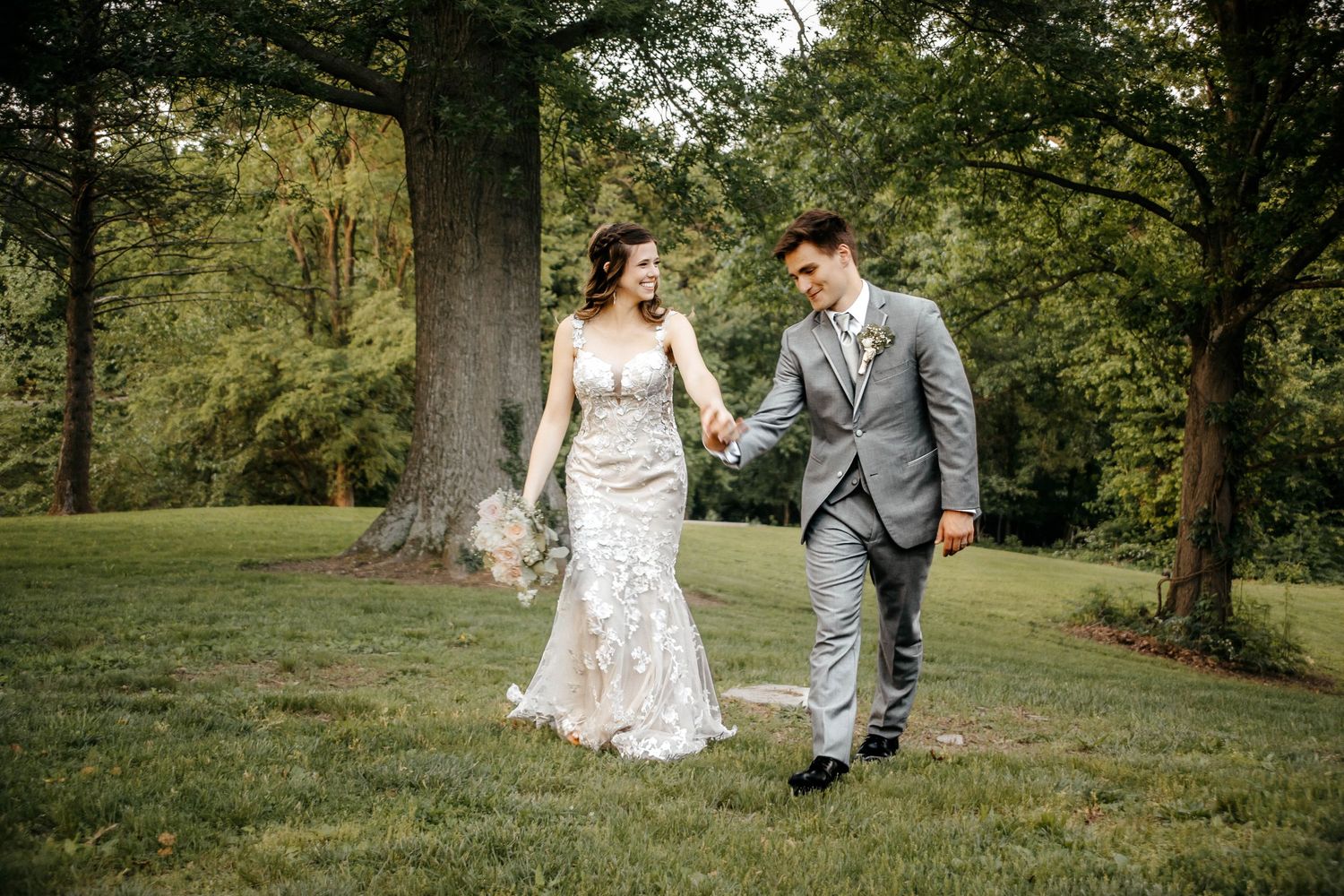 A bride and groom walk hand-in-hand on a lush green lawn surrounded by tall trees on their wedding day.