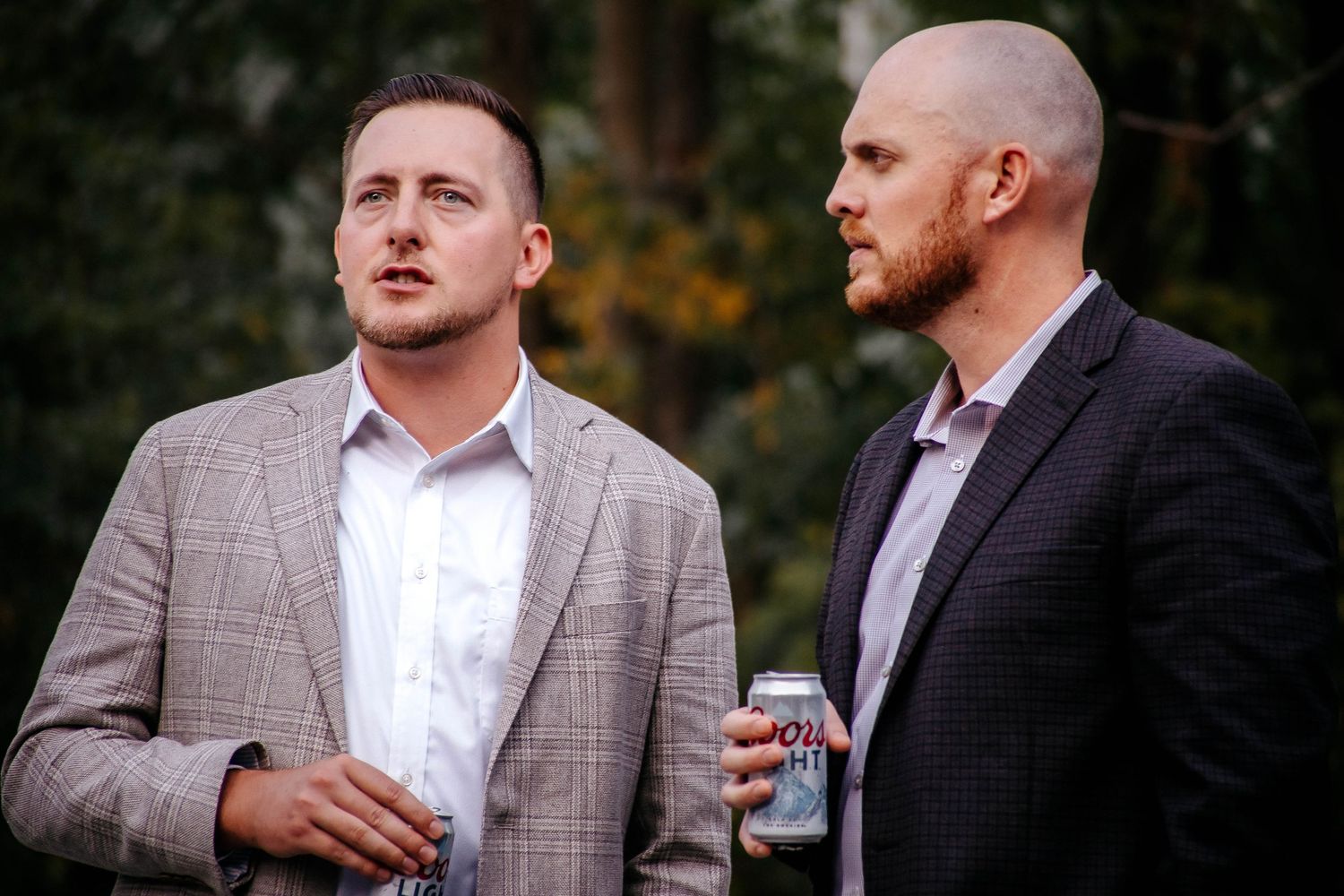 Two men in blazers stand outdoors, one holding a beer can, engaged in conversation at a social event.