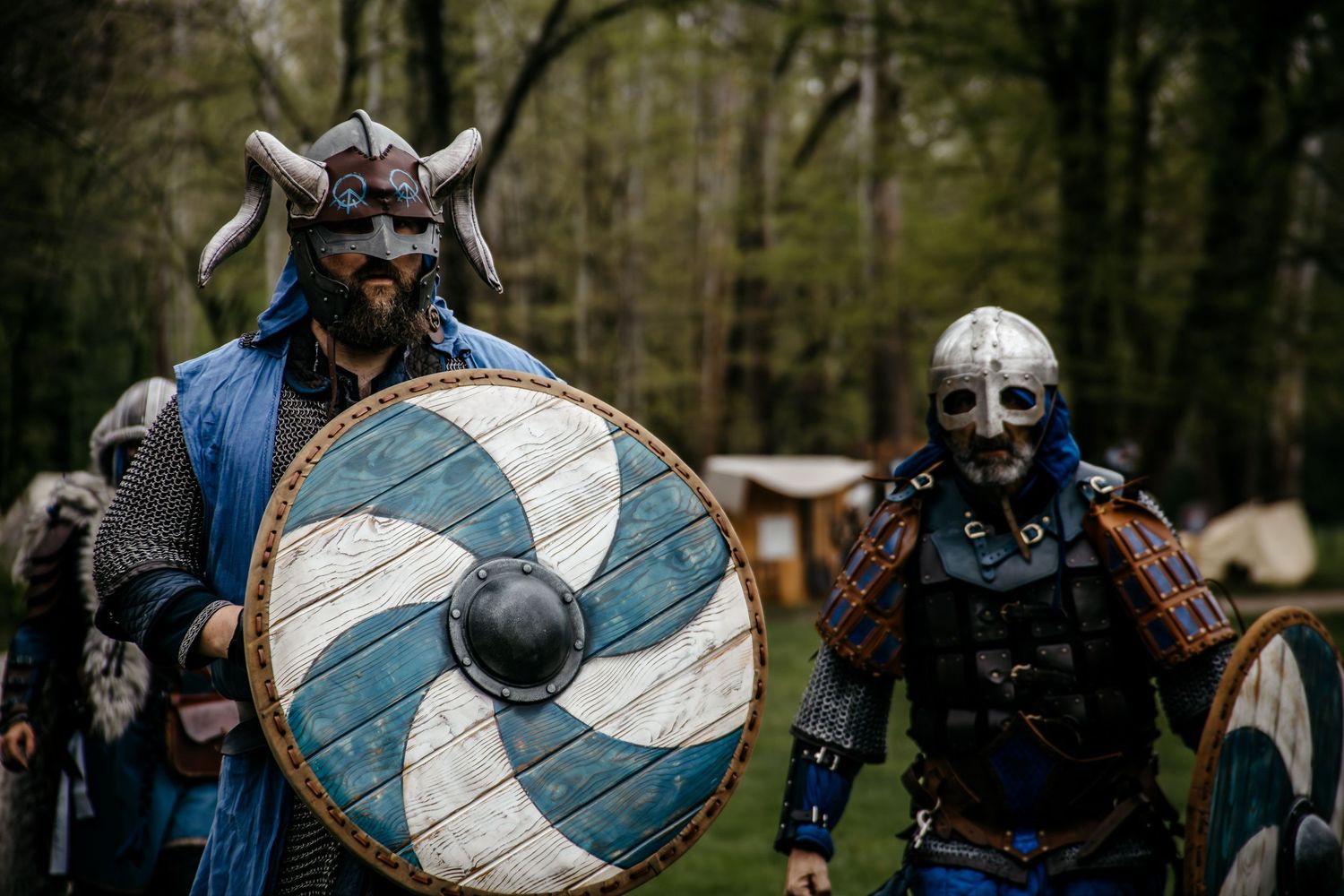 Two men dressed in Viking costumes with horned helmets and shields walk through a forest setting.