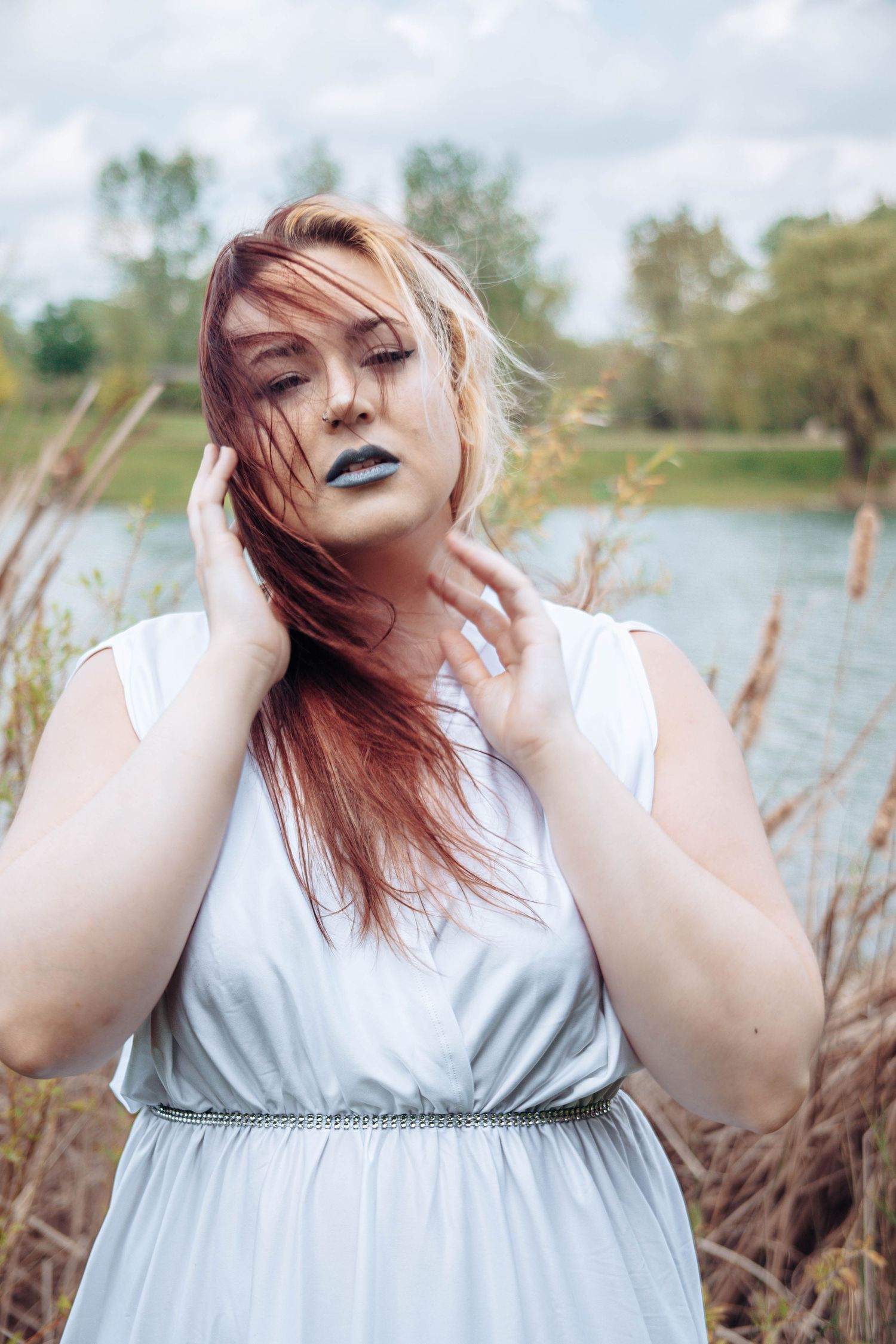 Red-haired woman in white dress with dark lipstick poses outdoors near a lake, hair blowing in the wind.