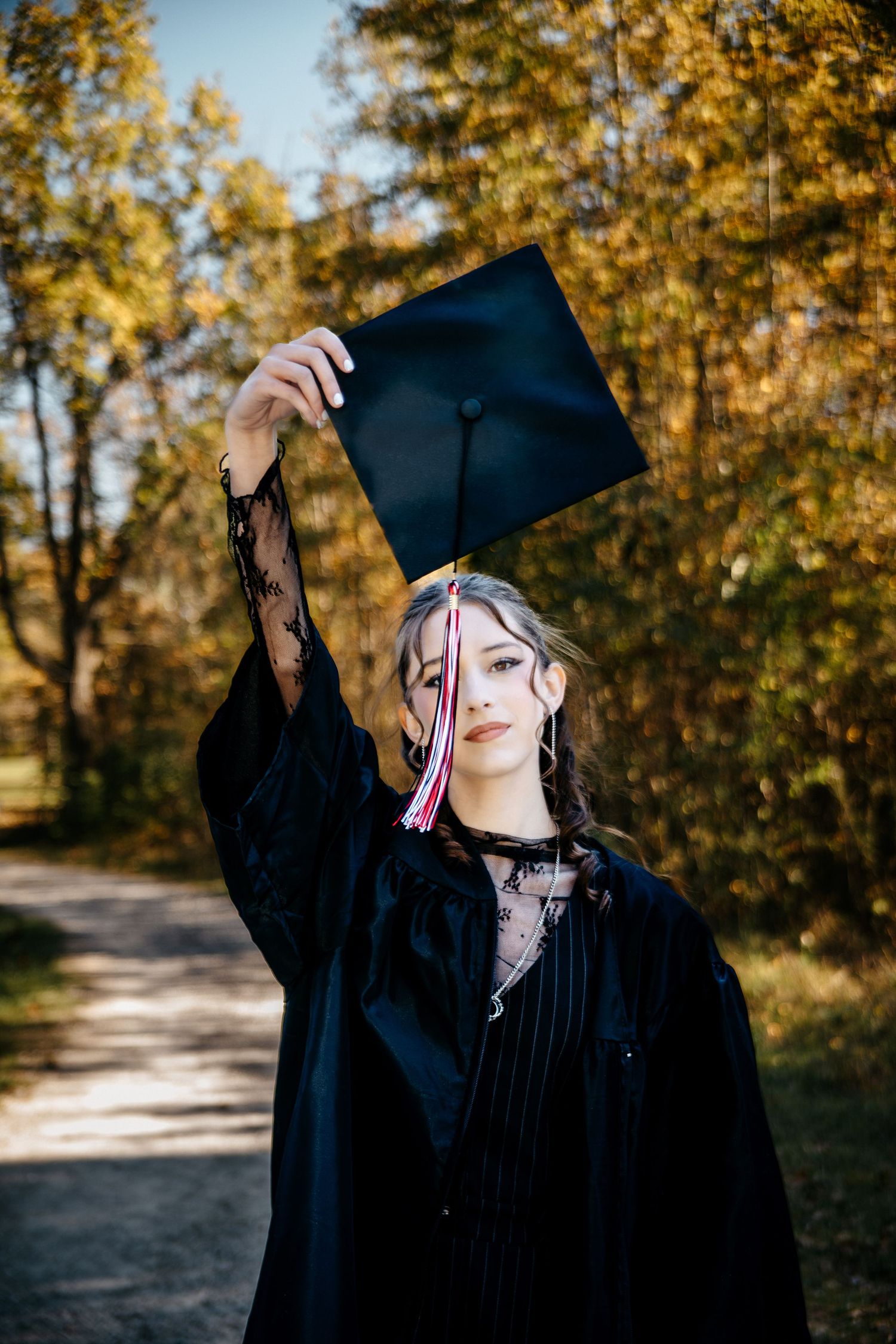 Graduate in black cap and gown celebrates achievement against autumn backdrop.