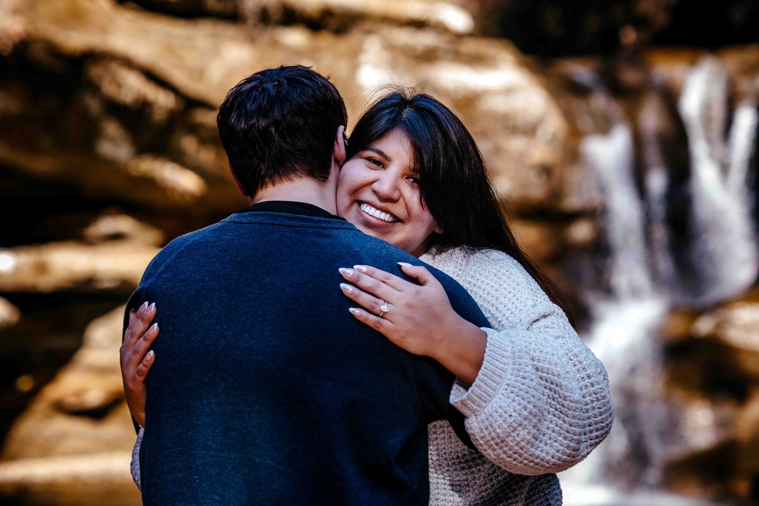 A joyful couple embraces near a waterfall, the woman smiling broadly in a cozy sweater while hugging her partner outdoors.