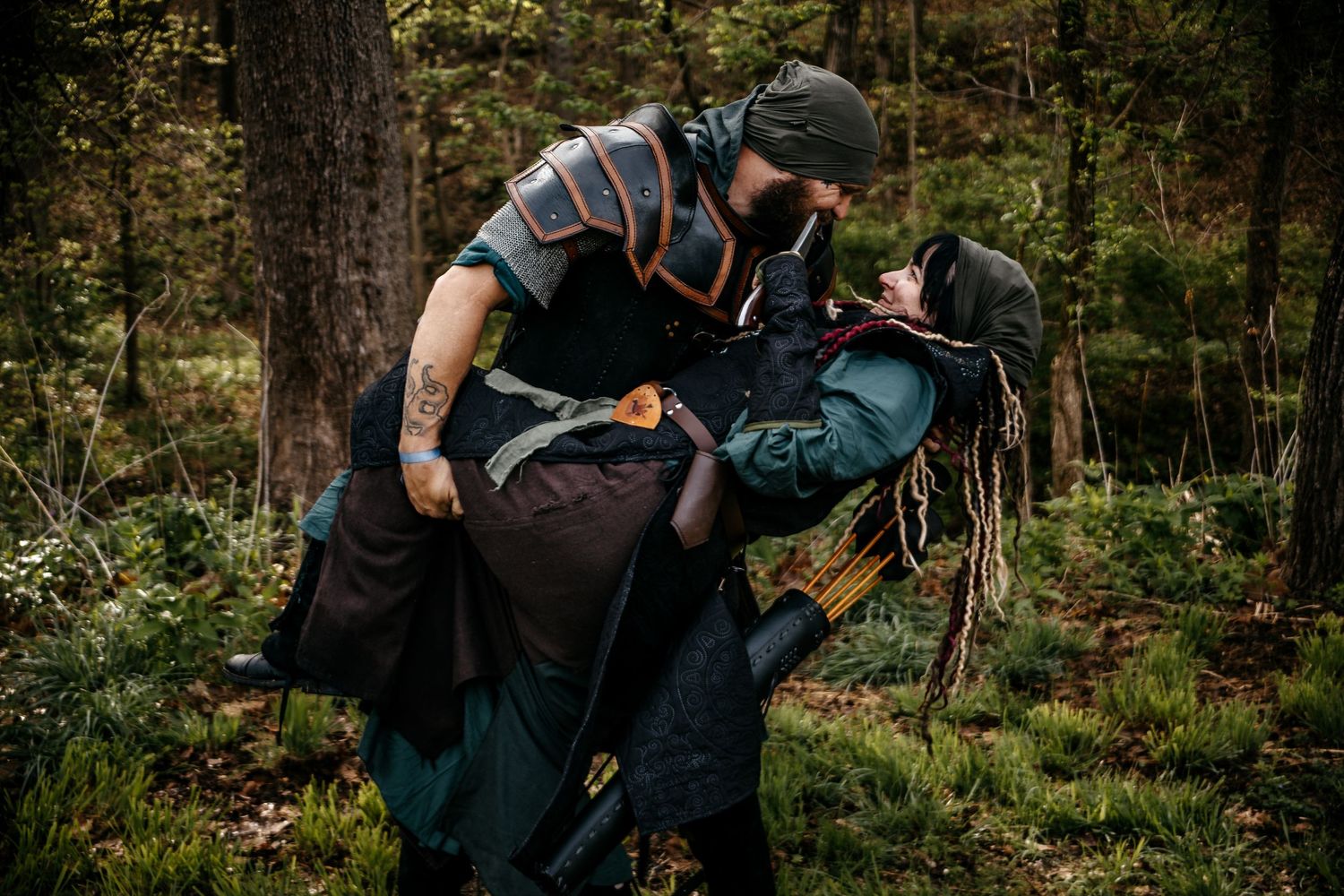 Two LARP cosplayers in medieval fantasy armor engage in combat roleplay in a forest setting.