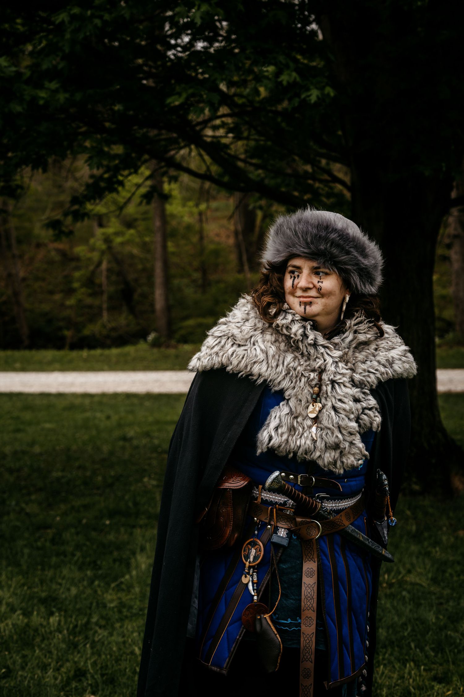 Person in medieval costume with fur hat, cape, and belt outdoors in a forest setting.