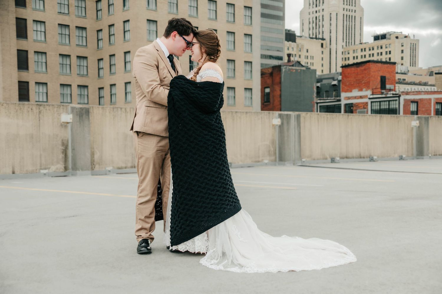 A couple in wedding attire share an intimate moment on an urban rooftop with city buildings in the background.