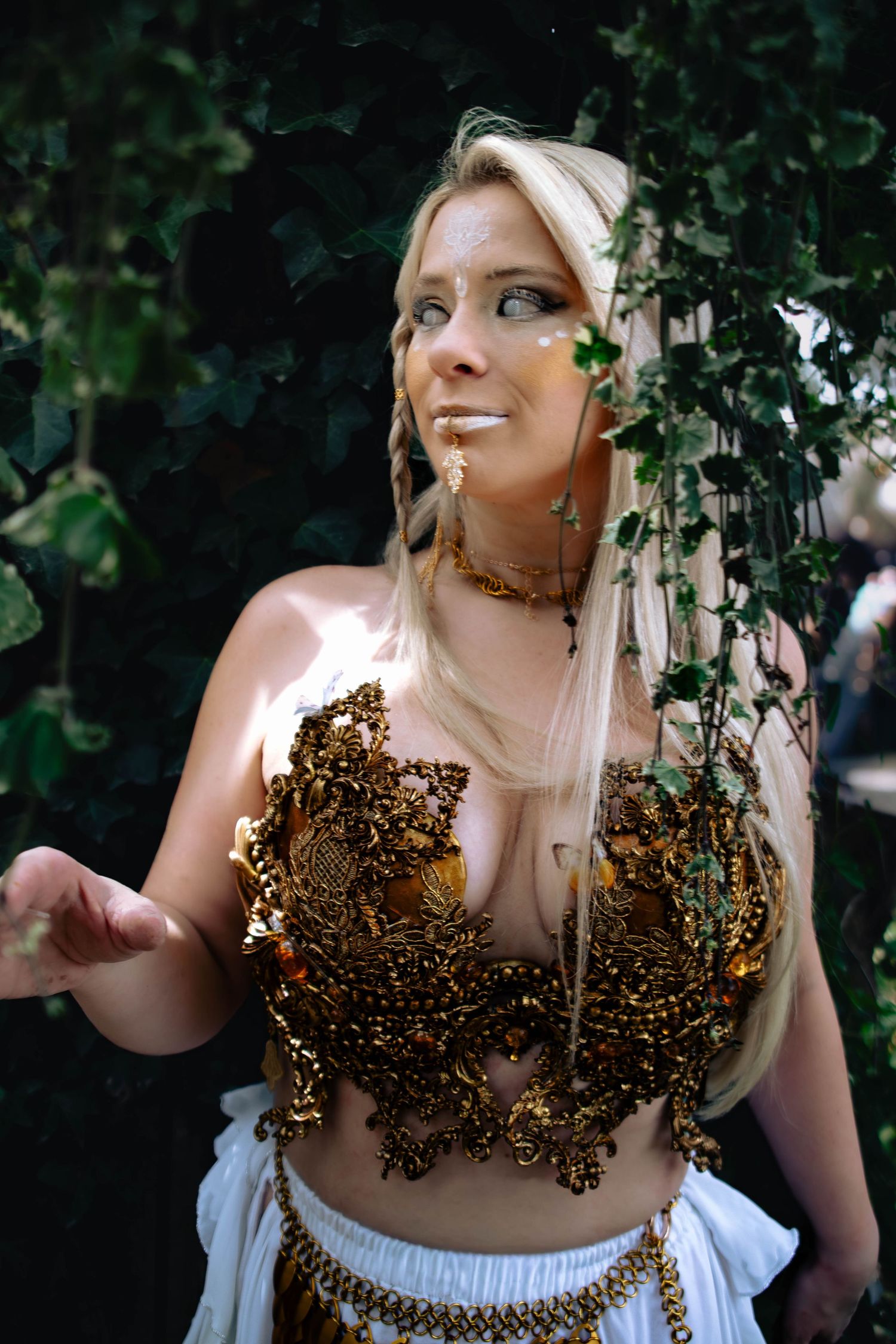 Blonde woman in ornate gold costume with ivy backdrop at outdoor festival.
