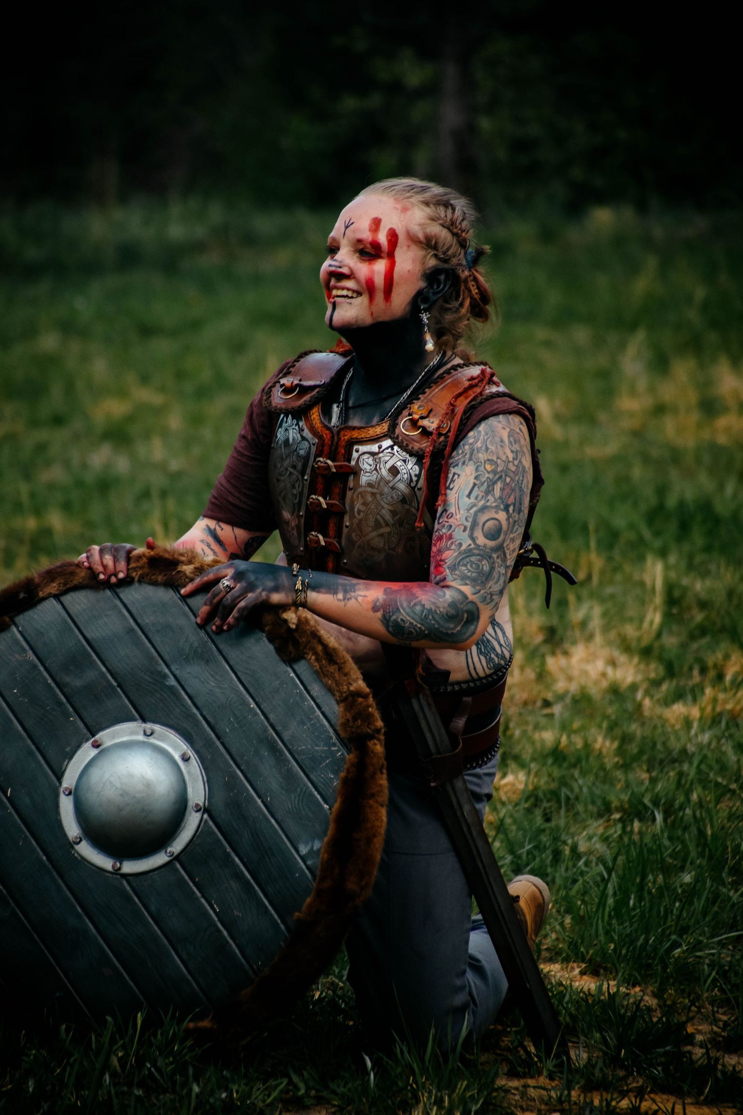 A warrior with face paint and armor poses with a round shield on grassy ground.