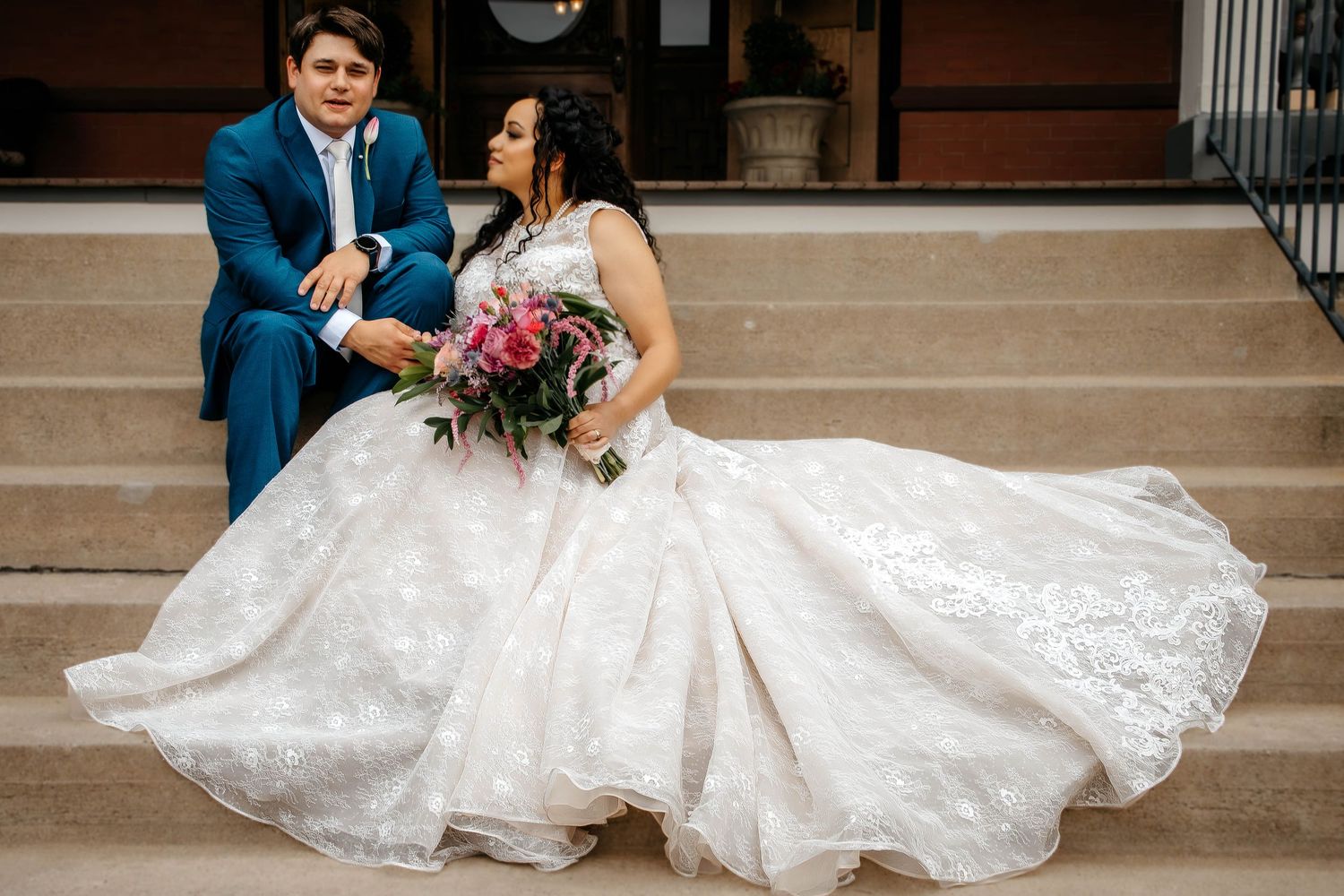 A romantic couple sits on stone steps with the bride's white lace dress elegantly spread around them.