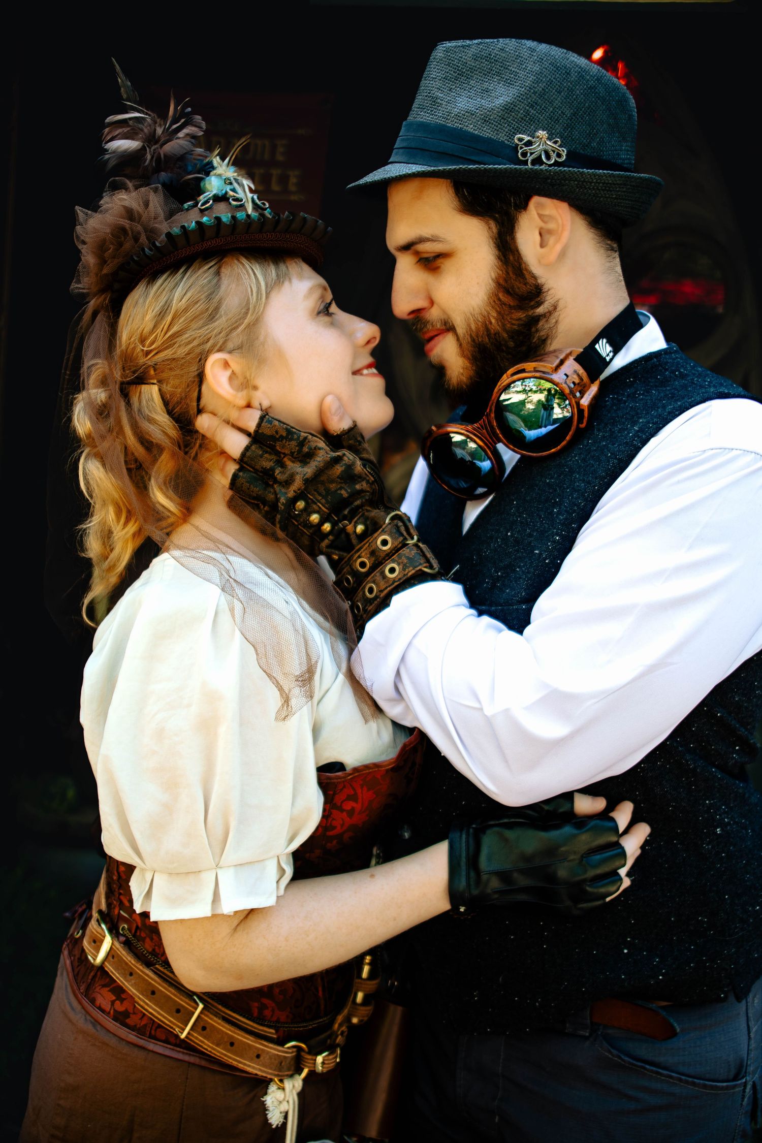 A romantic steampunk-themed couple embraces in vintage Victorian-inspired costumes against a dark background.
