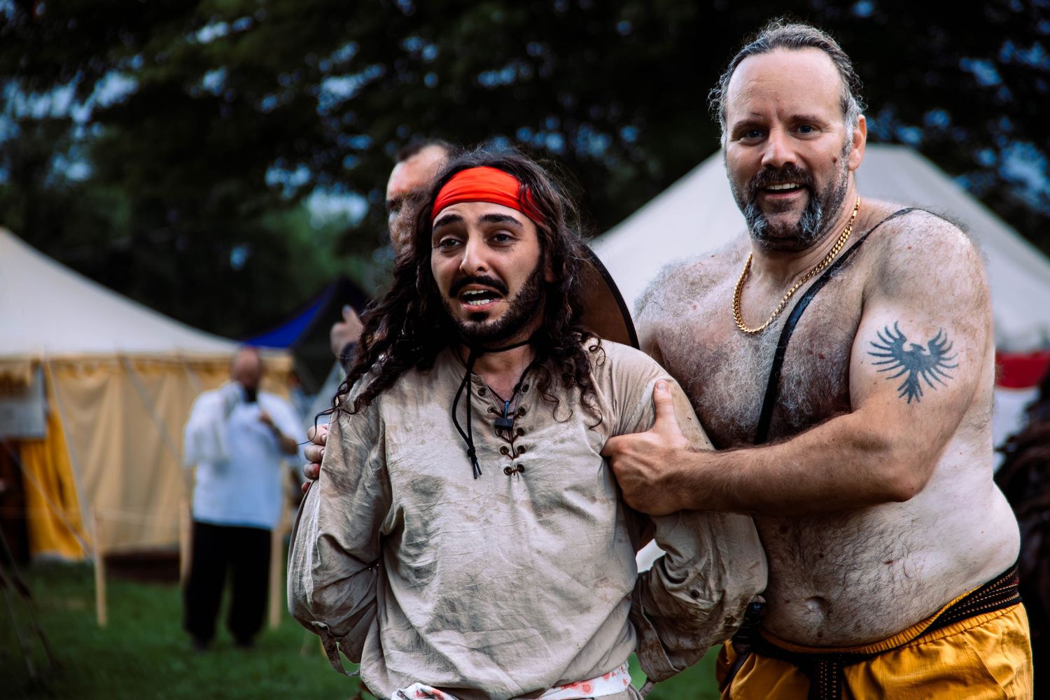 Two performers at a medieval festival share a laugh together near canvas tents in the background.