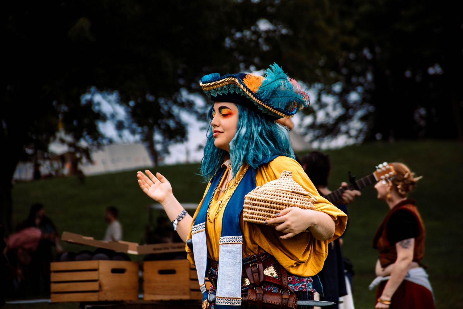 A performer in a blue wig and yellow jacket sings on stage during an outdoor festival performance.