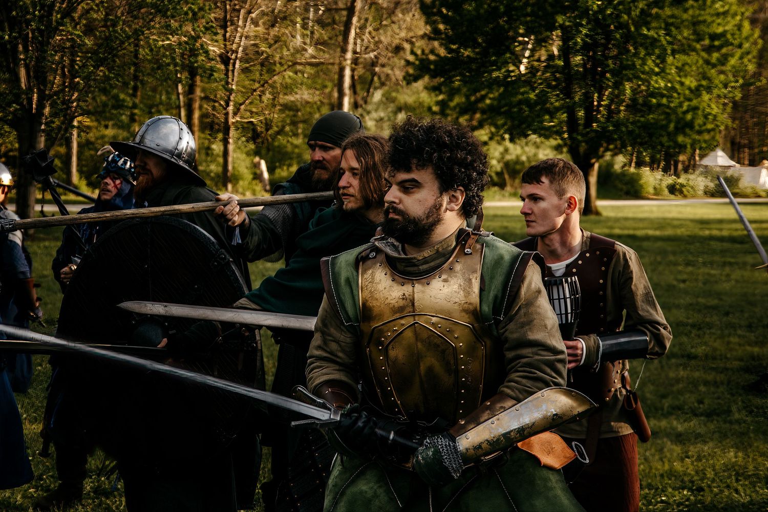 Medieval reenactors in armor wielding spears and swords during an outdoor historical battle demonstration in a park.