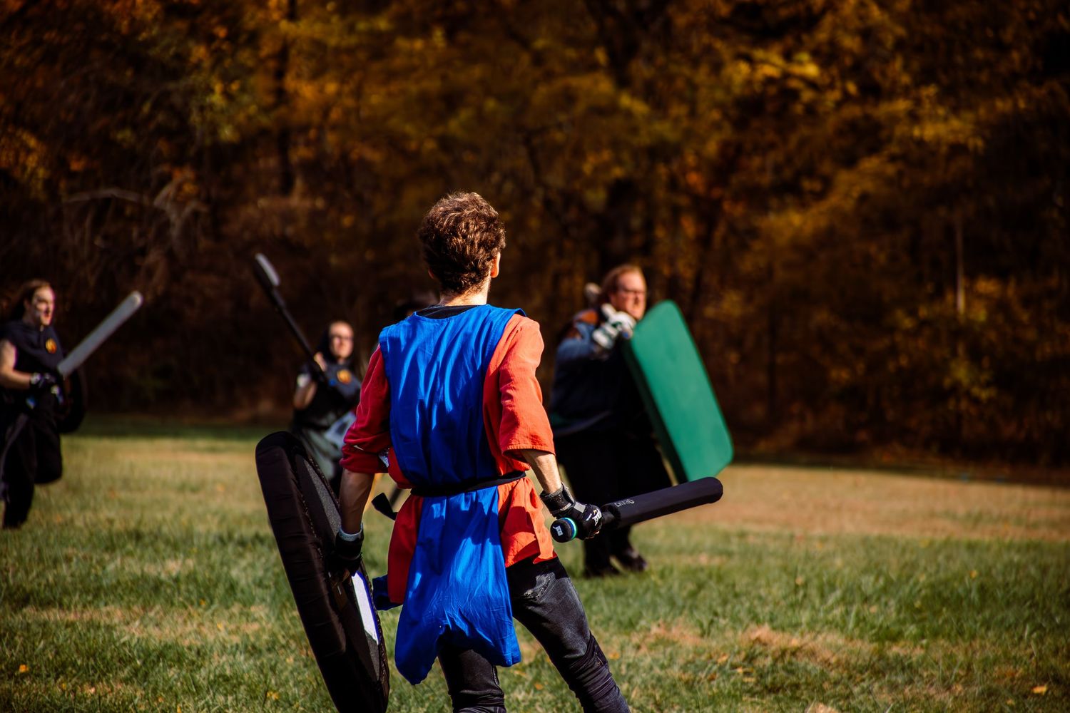 Medieval LARP battle reenactment with costumed participants wielding shields and weapons in an autumn park setting.