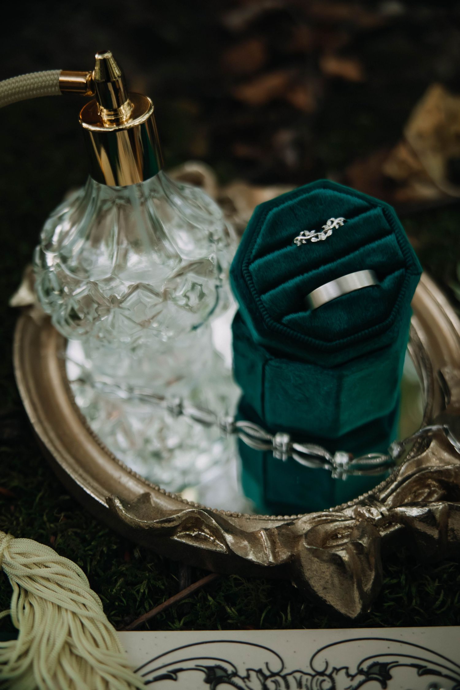 A vintage glass perfume bottle and emerald velvet ring box with wedding rings displayed on an ornate silver tray.