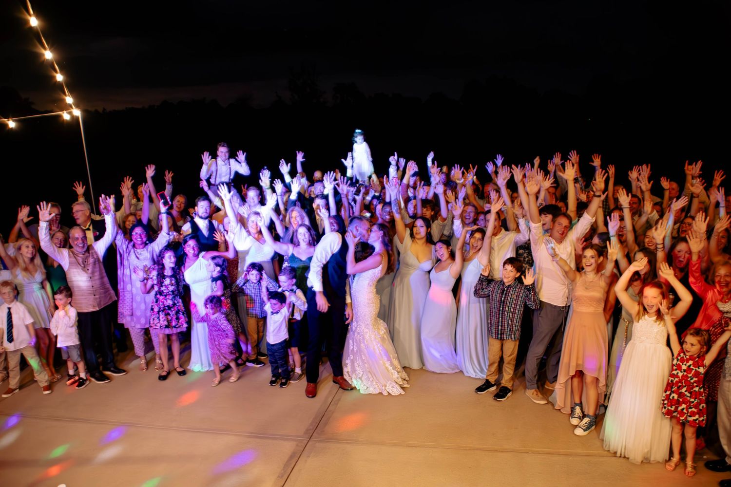 Large wedding party and guests celebrate on outdoor dance floor at night with colorful lights and hands raised.