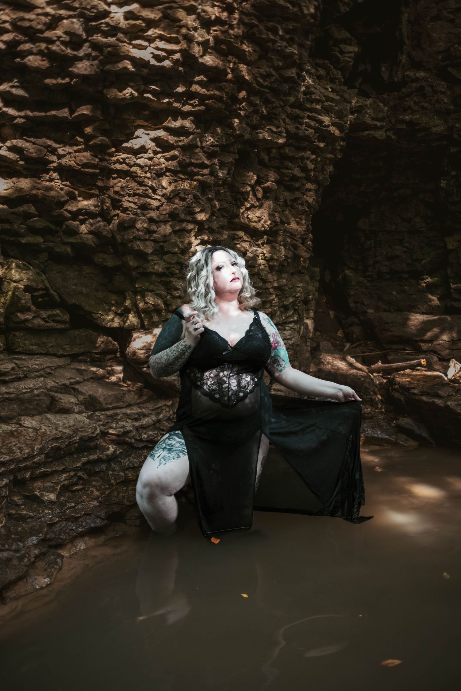 Moody boudoir photography in black dress against rocky cave wall and shallow water.