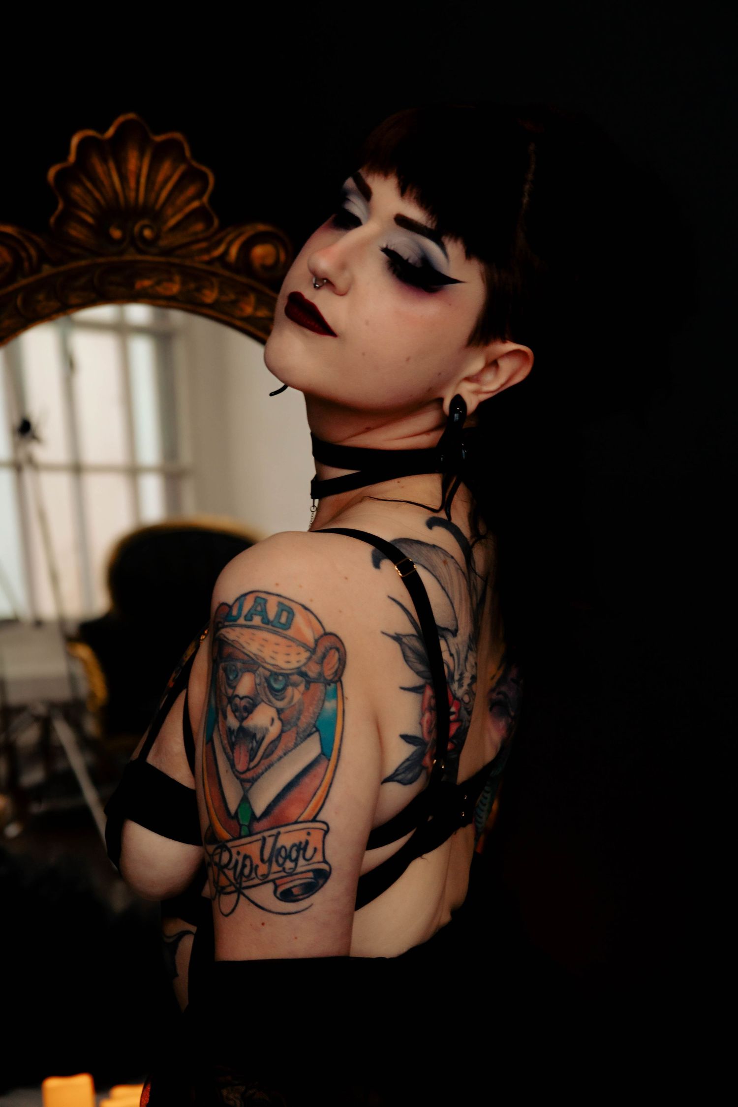 Artistic portrait showing detailed tattoo art and gothic styling with ornate mirror in background.