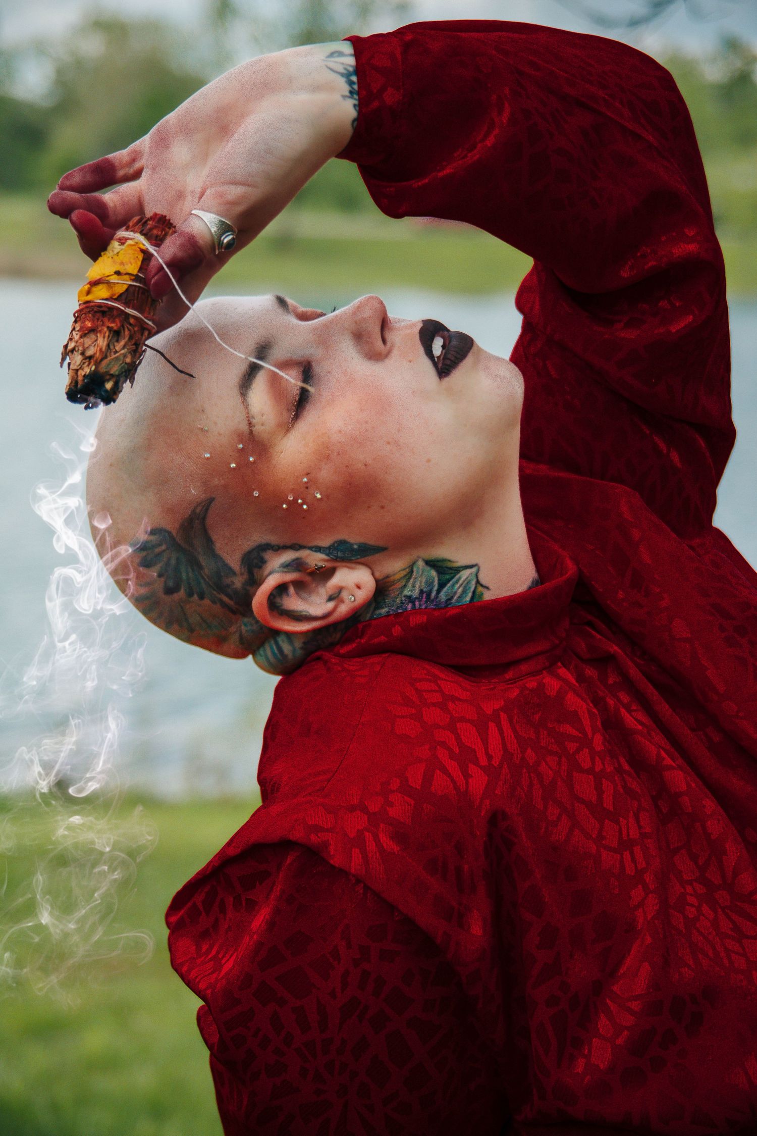 Person in red velvet top exhales smoke while wearing tattoos and holding an amber crystal in artistic portrait.