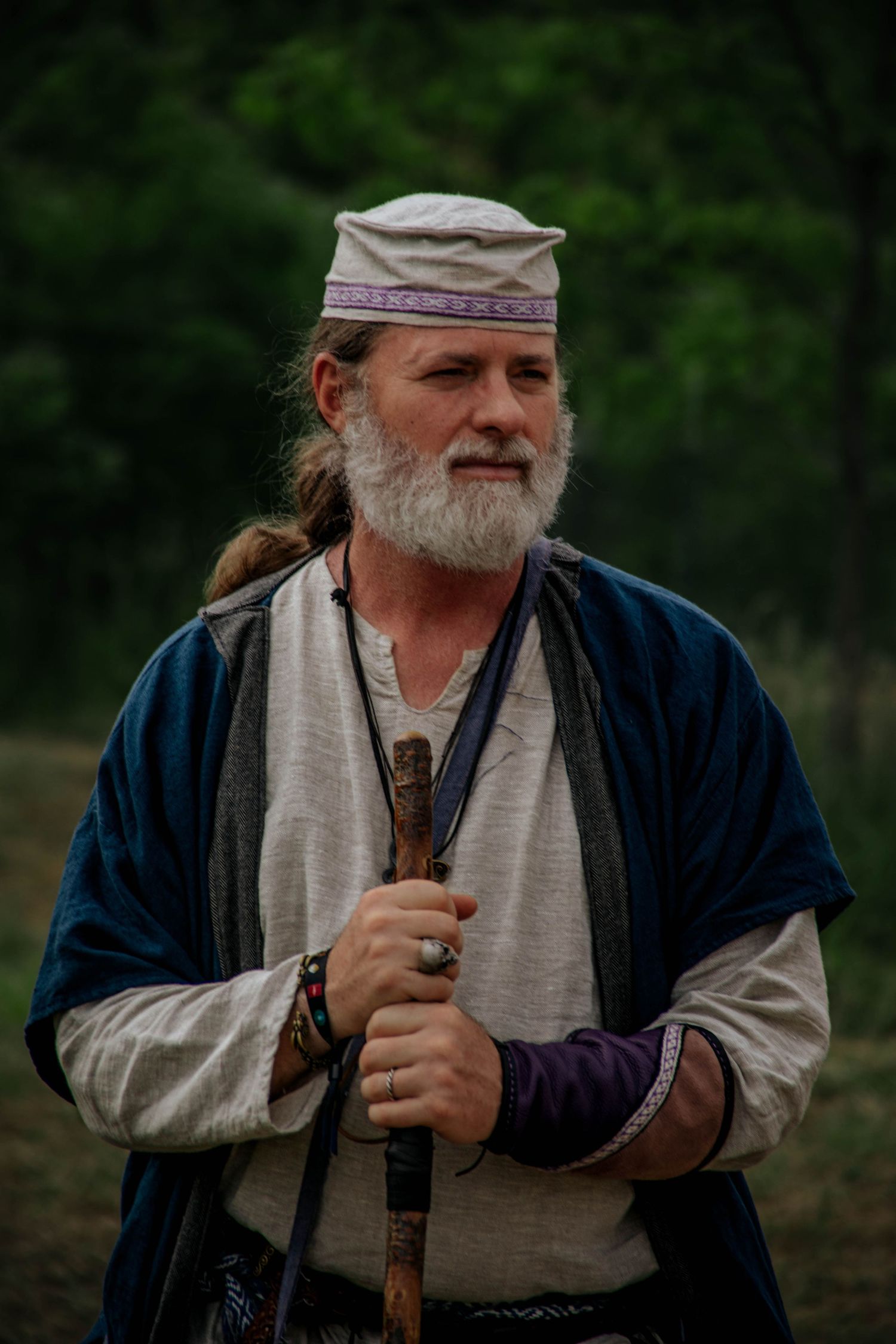 A person in medieval period clothing holds a wooden staff while wearing a white scarf and navy tunic.