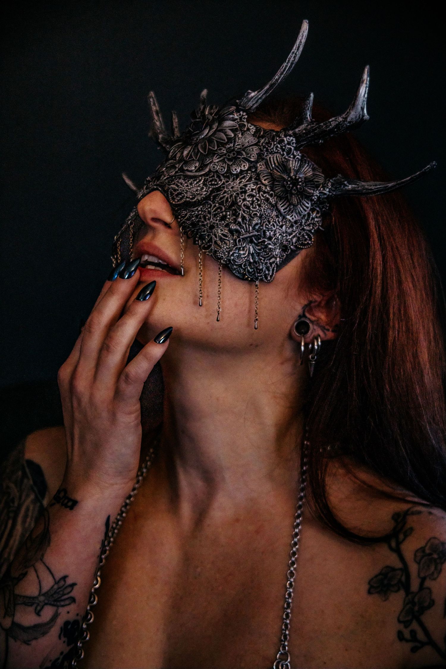 Woman wearing ornate dragon mask with antlers, dark nails, tattoos, and chain jewelry poses dramatically against dark background.