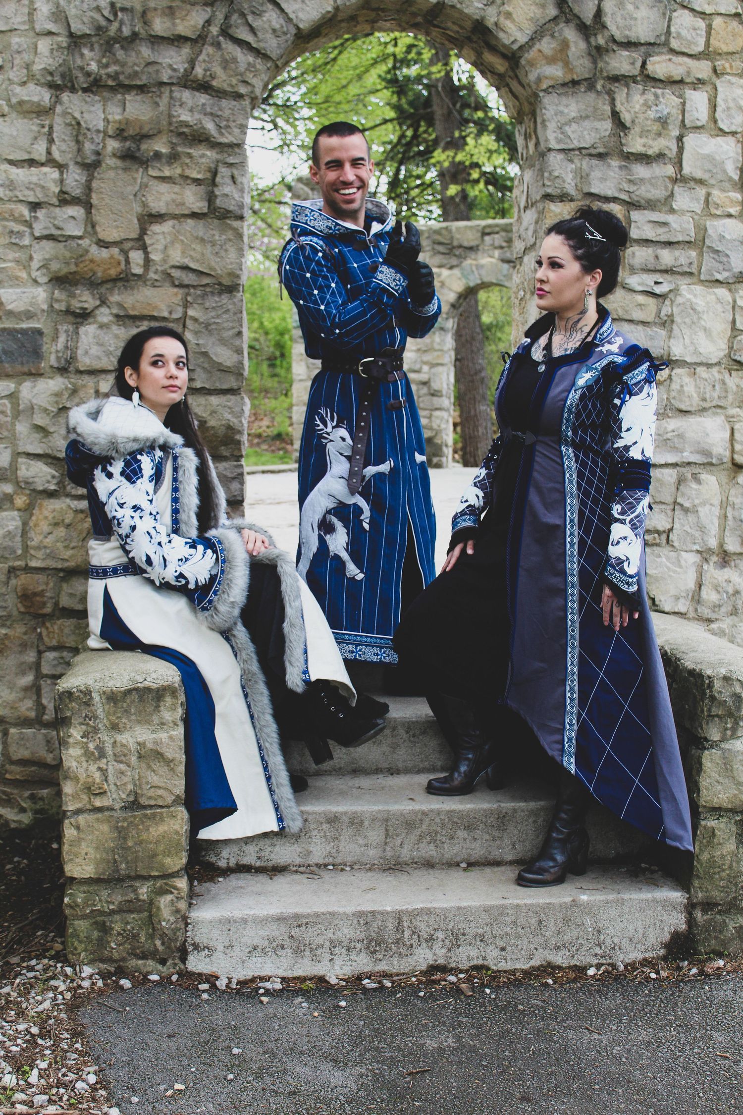 Three cosplayers in medieval fantasy costumes with blue and white designs pose in a stone archway.