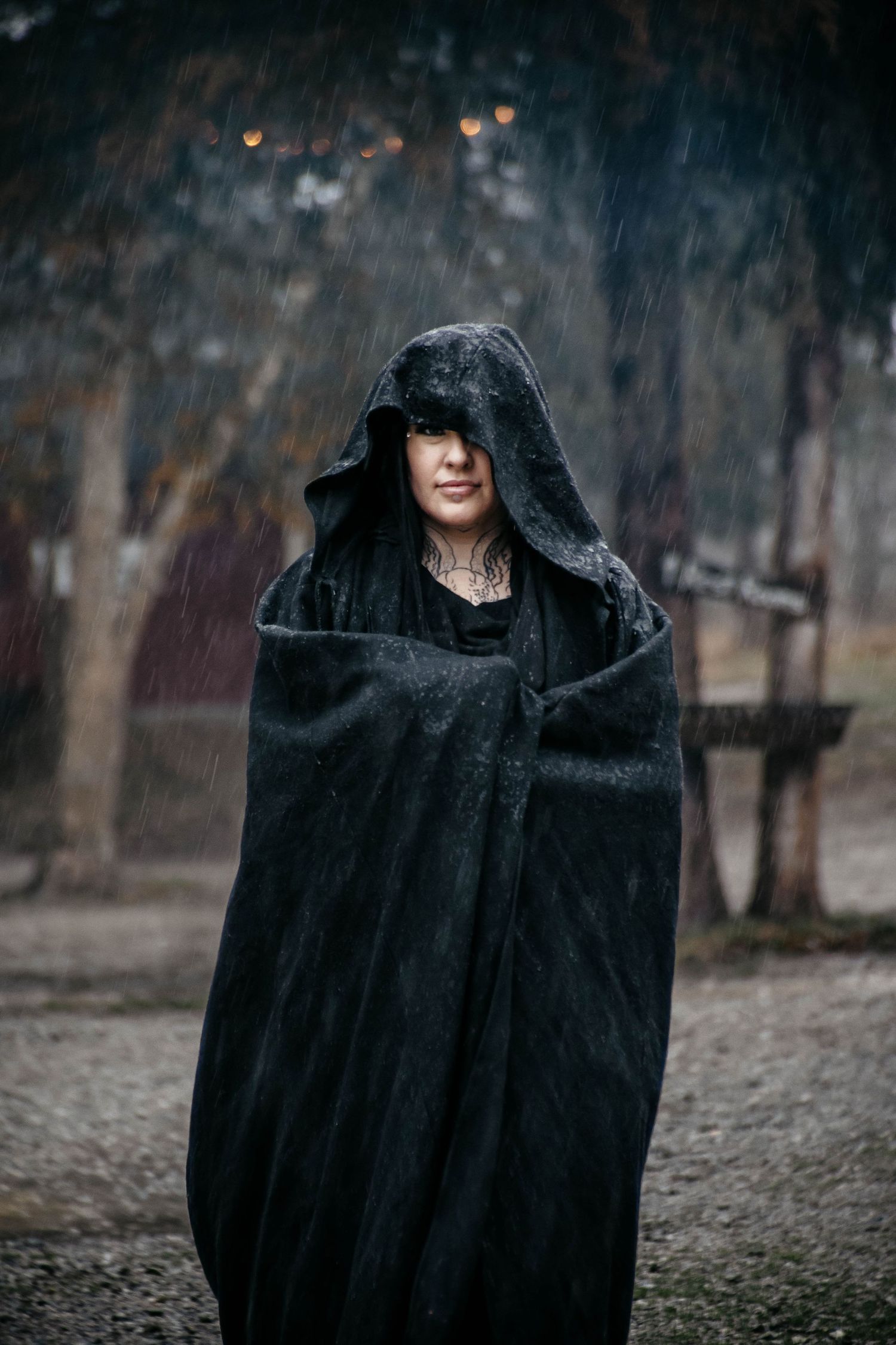 A mysterious figure in a black hooded cloak stands in a moody, dark outdoor setting.