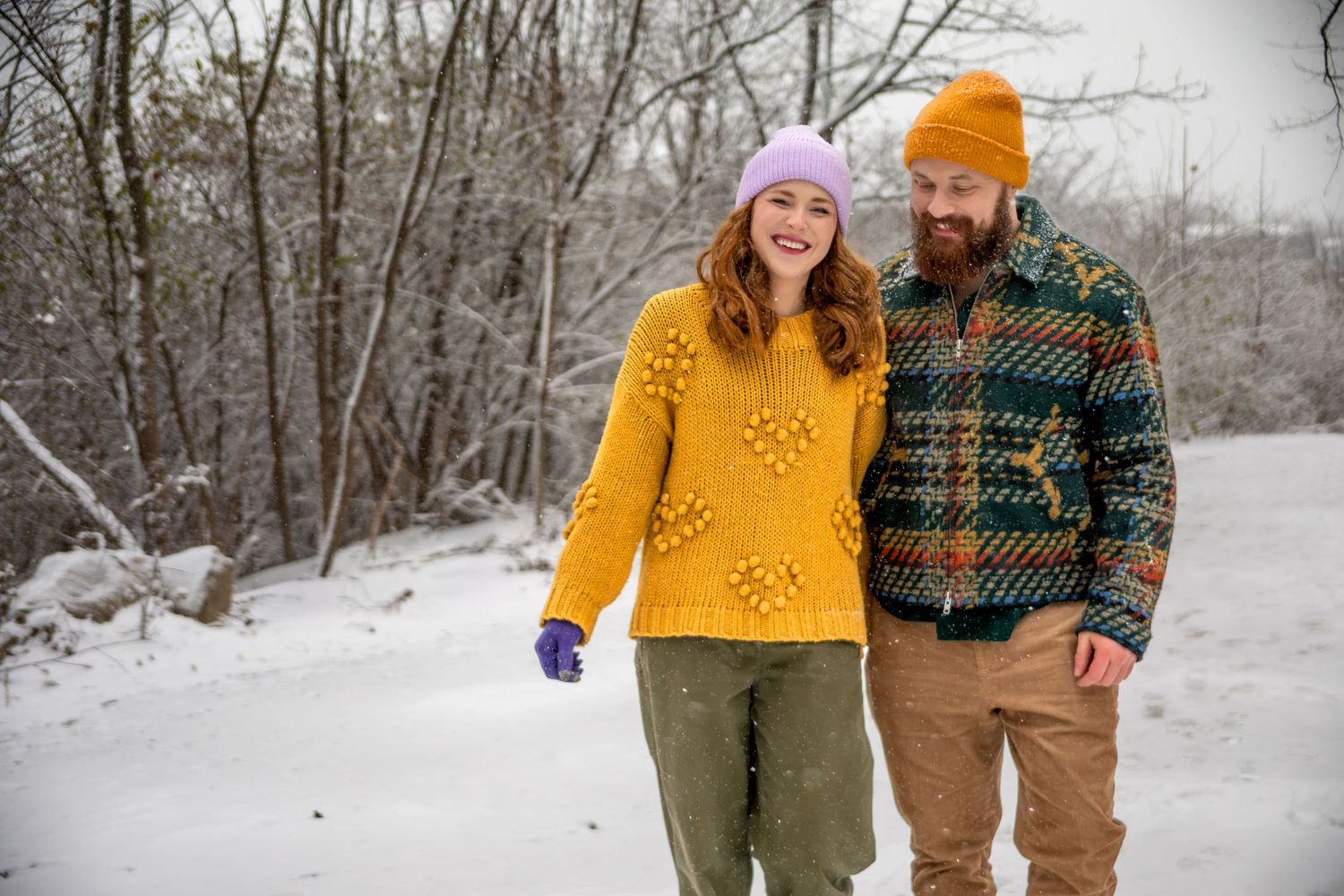 A couple wearing winter clothing walks through a snowy forest landscape holding hands.