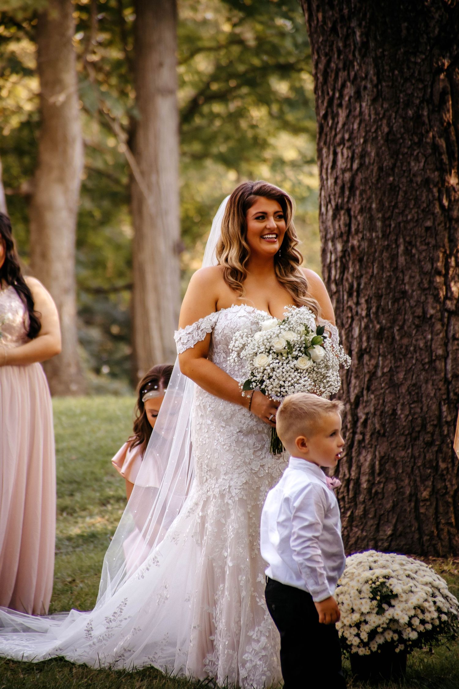 A beautiful outdoor wedding scene with a bride in a lace off-shoulder gown holding white flowers in a wooded setting.