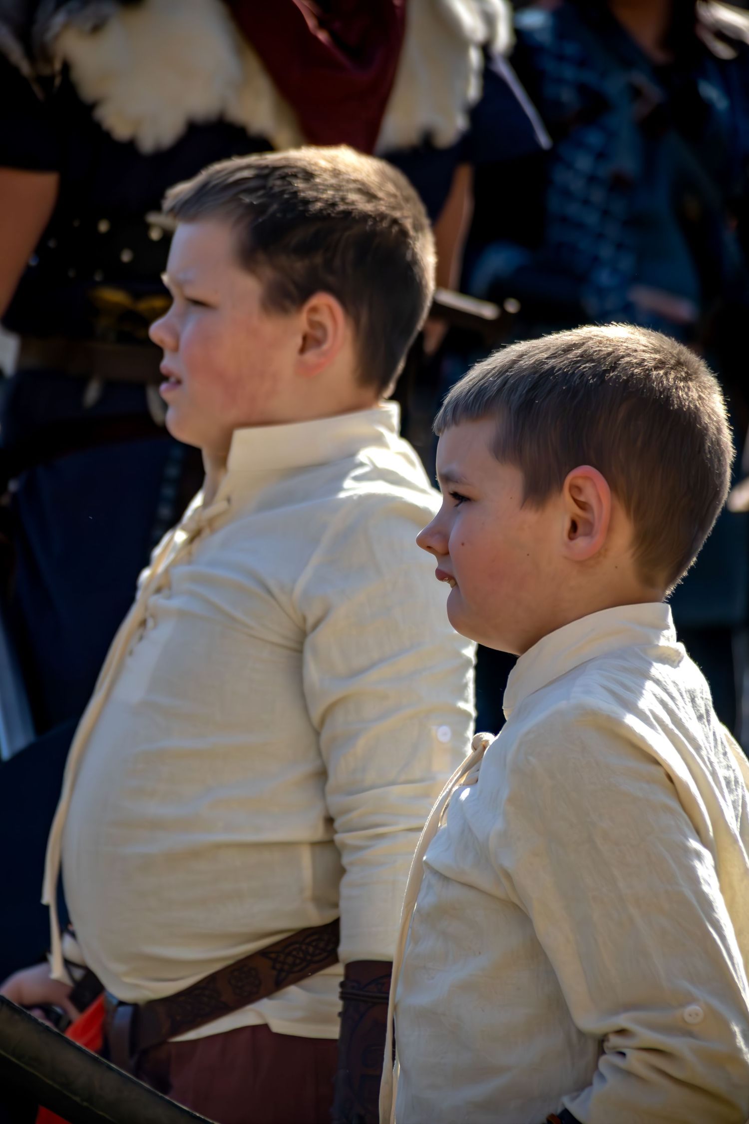Two young boys in matching cream-colored shirts sit together at an outdoor event in natural sunlight.