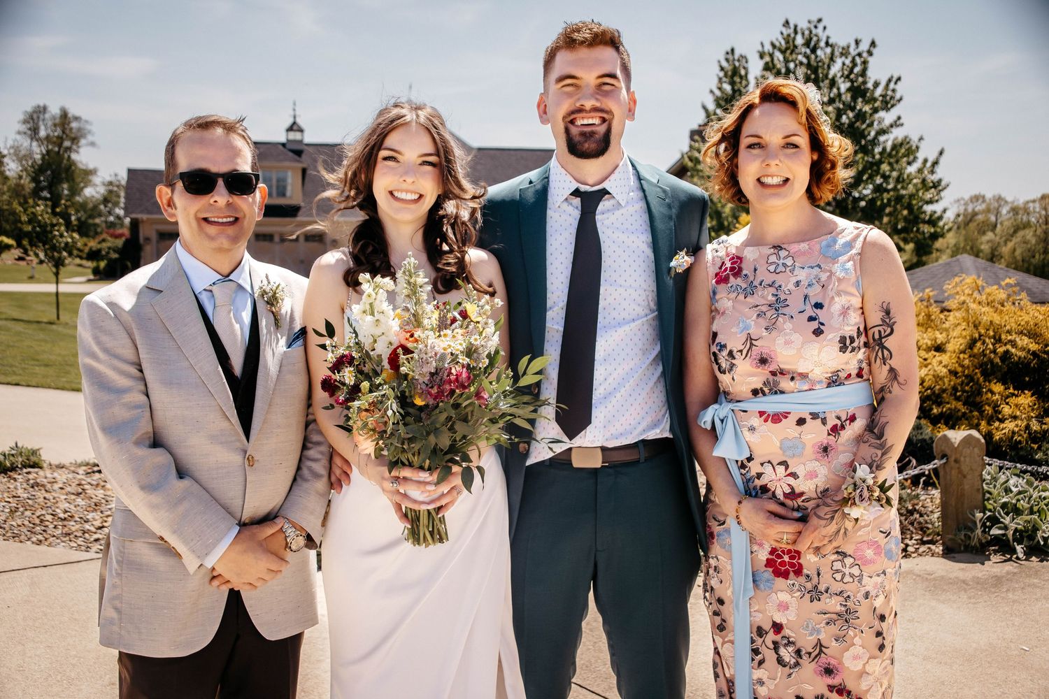 Wedding party poses outdoors in casual attire with floral bouquets against a sunny backdrop.