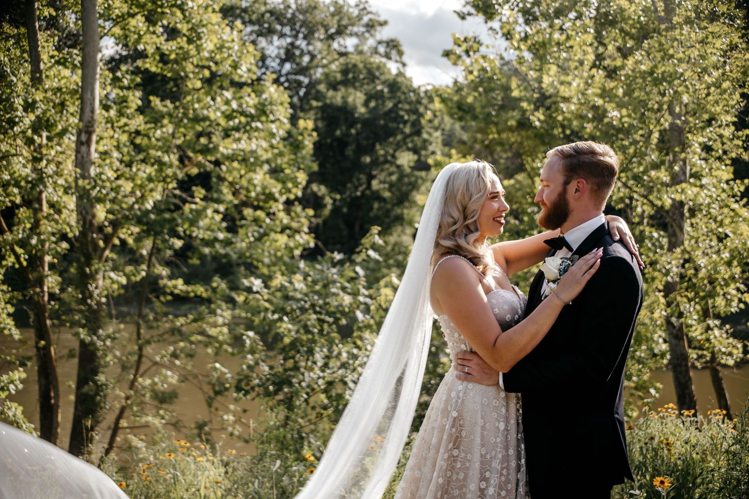 Couple embracing in an outdoor garden setting with lush green trees in the background.