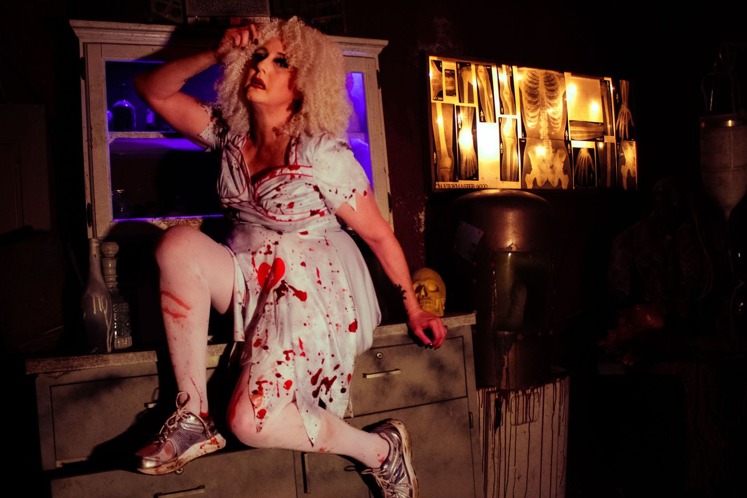 Woman in blood-splattered white dress poses dramatically in dark haunted house Halloween scene with eerie lighting.