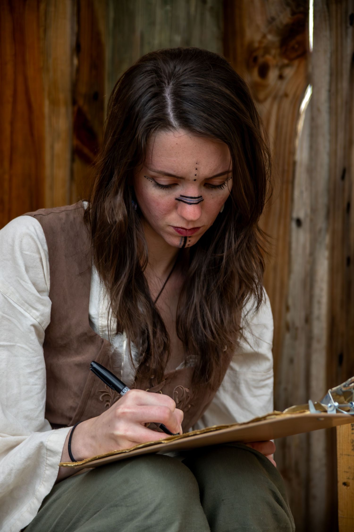 Young woman with face paint and brown hair writes on clipboard, wearing white blouse and brown vest near wooden wall.