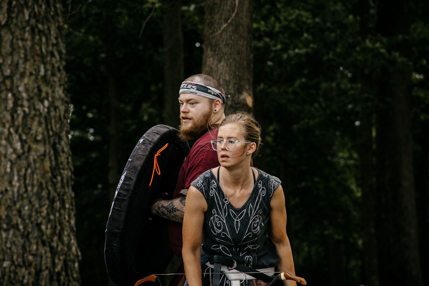 Two athletes rest in a forest setting, man carrying a tire, woman in sleeveless top, during an obstacle race event.