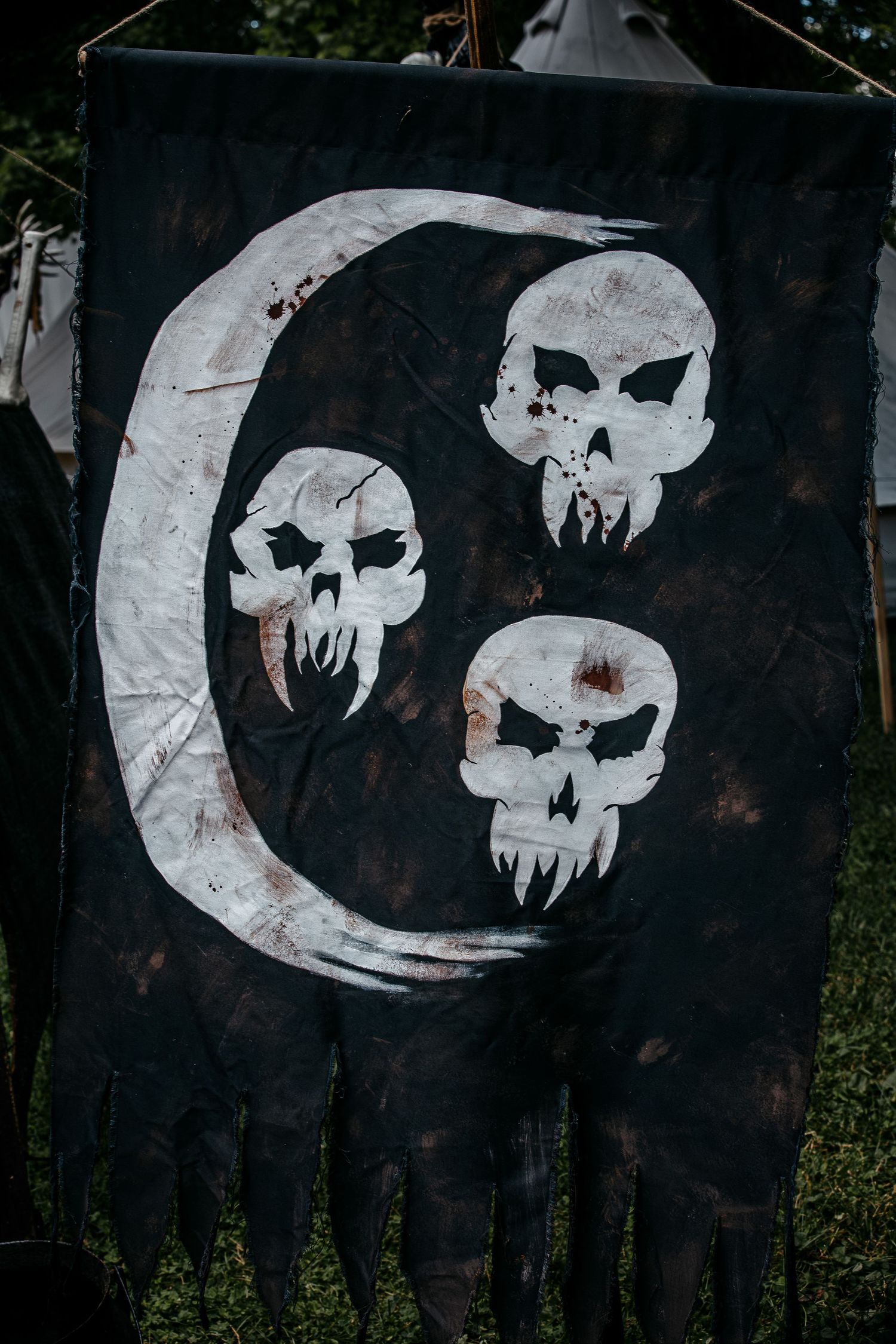 Dark medieval banner with white crescent moon and three skull designs, displayed outdoors at a fantasy or LARP event.