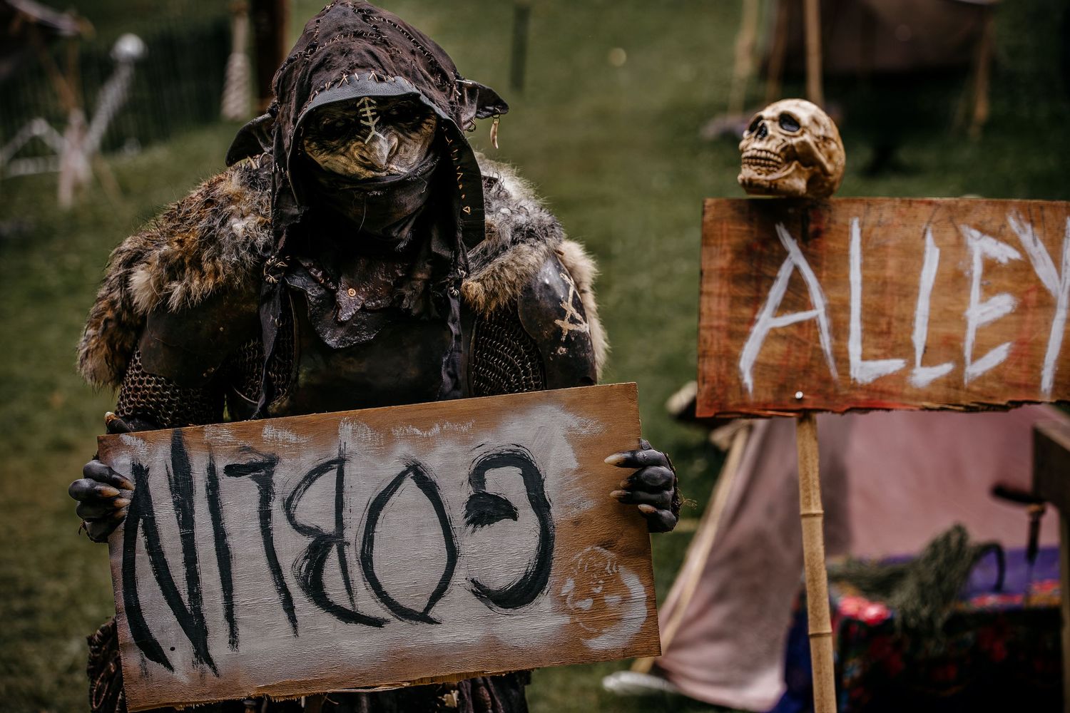 A costumed goblin character holds a sign reading Goblin Alley at a fantasy themed outdoor event.