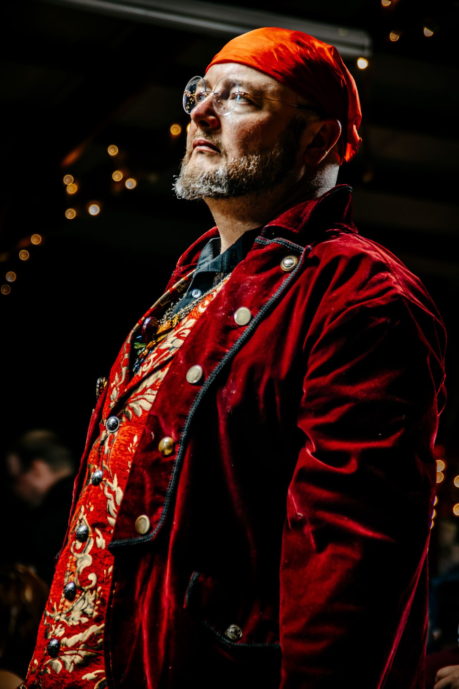 Man in red velvet coat, orange bandana, and glasses gazes upward at a festive event with bokeh lights.