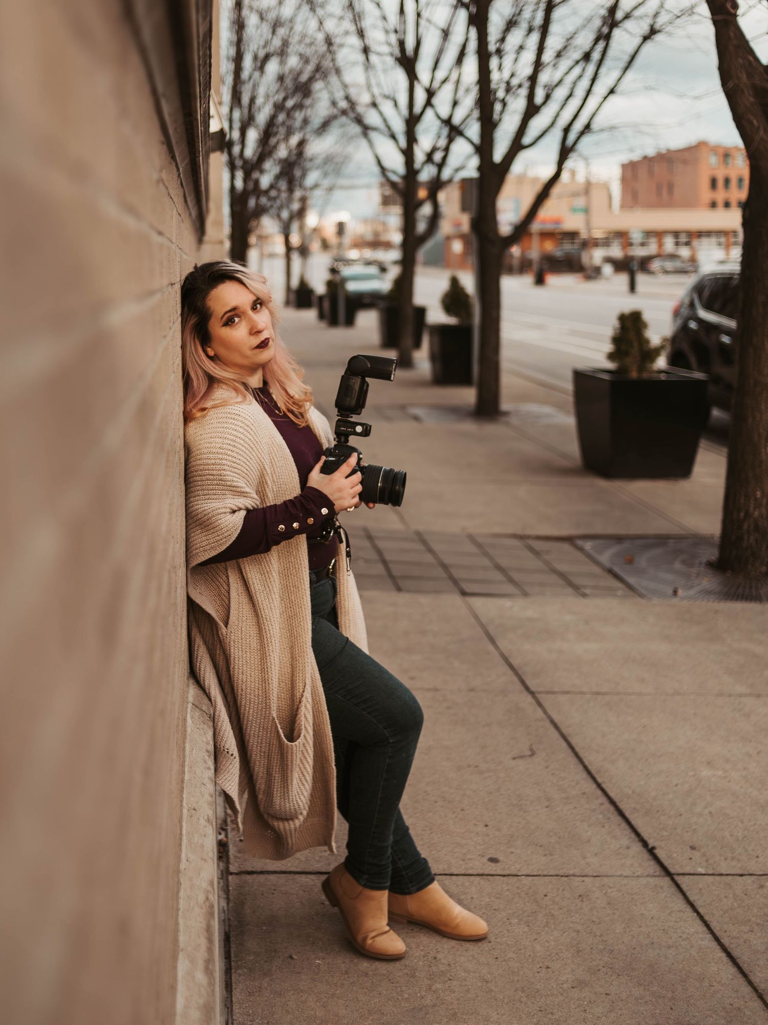 Urban portrait of photographer with camera leaning against building wall on city sidewalk.