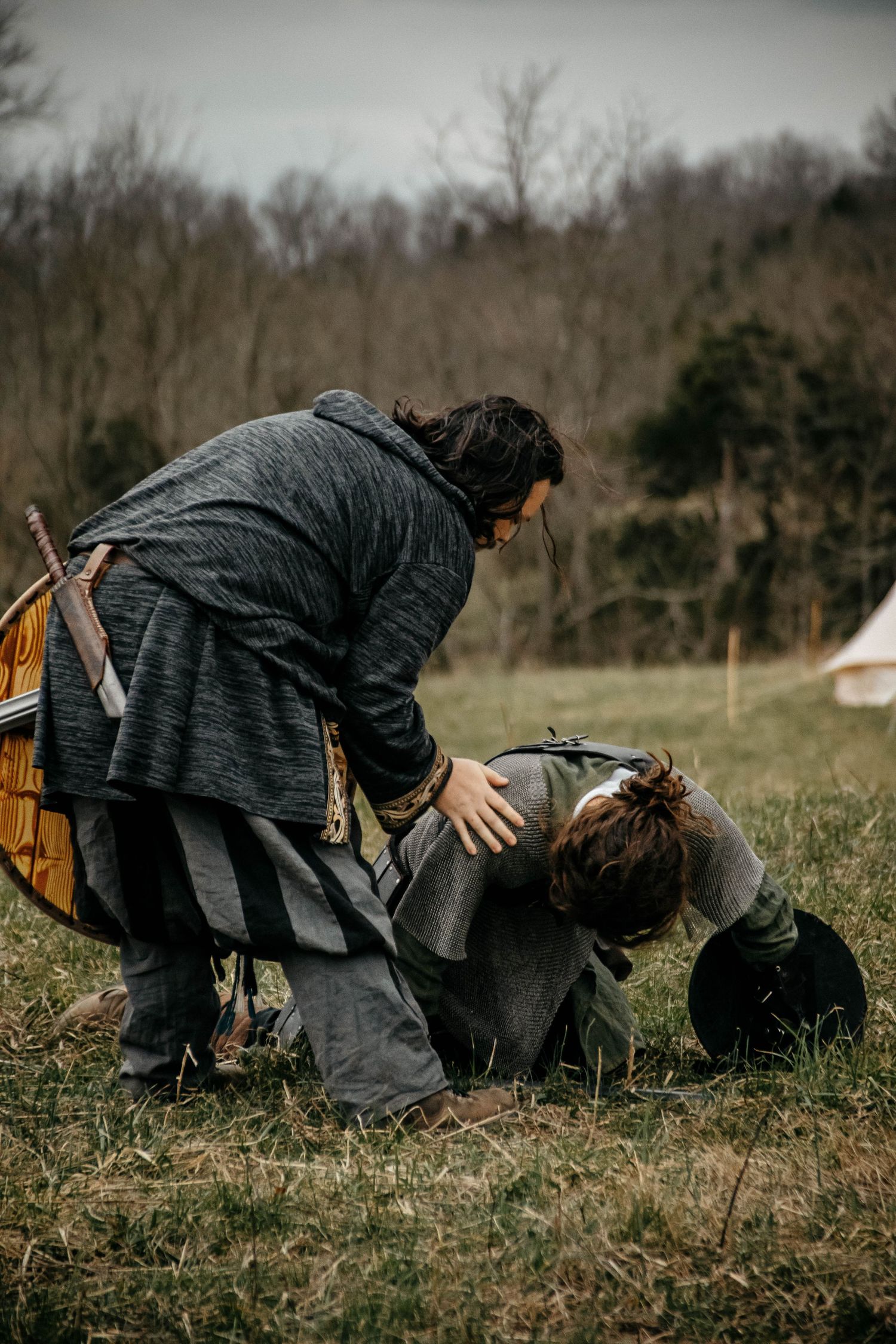 Two people in medieval or historical costumes interact on a grassy field with bare trees in the background.