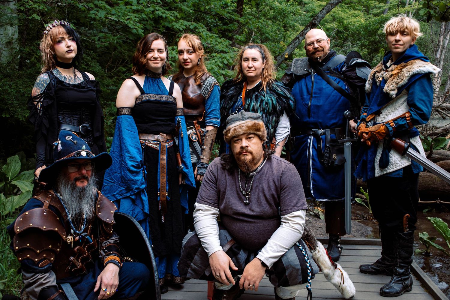 A group of medieval cosplay enthusiasts in period costumes gather outdoors in a forest setting.