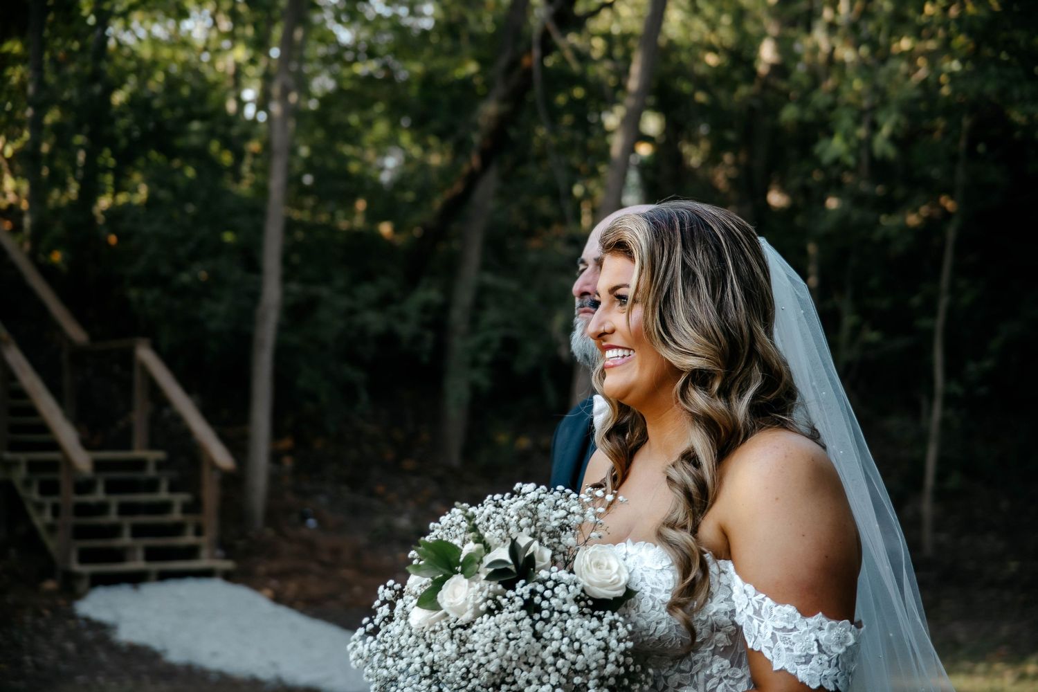 A joyful bride in a white off-shoulder gown laughs during an outdoor wedding photoshoot in a wooded setting.