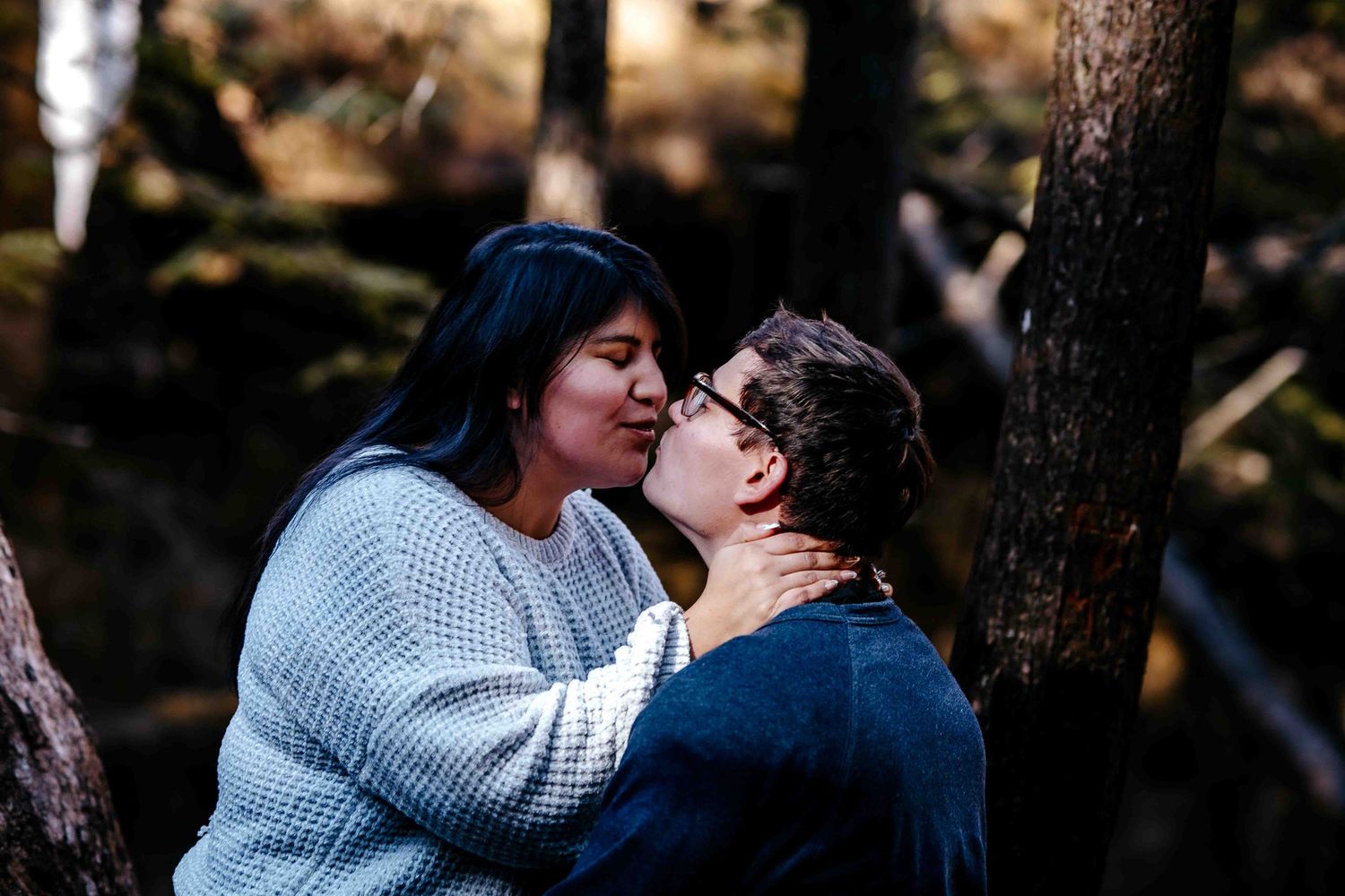 Couple sharing a romantic kiss outdoors in a forest setting, woman in blue sweater embracing man near trees.