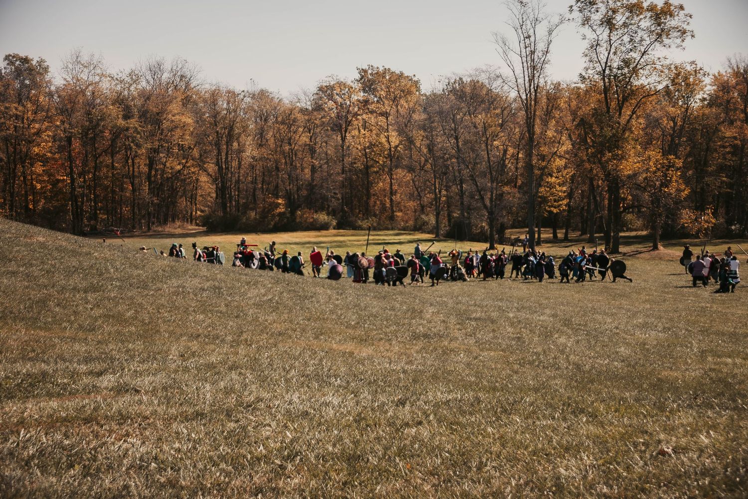 A panoramic view of a grassy field with autumn trees and people gathered in the distance during fall season.