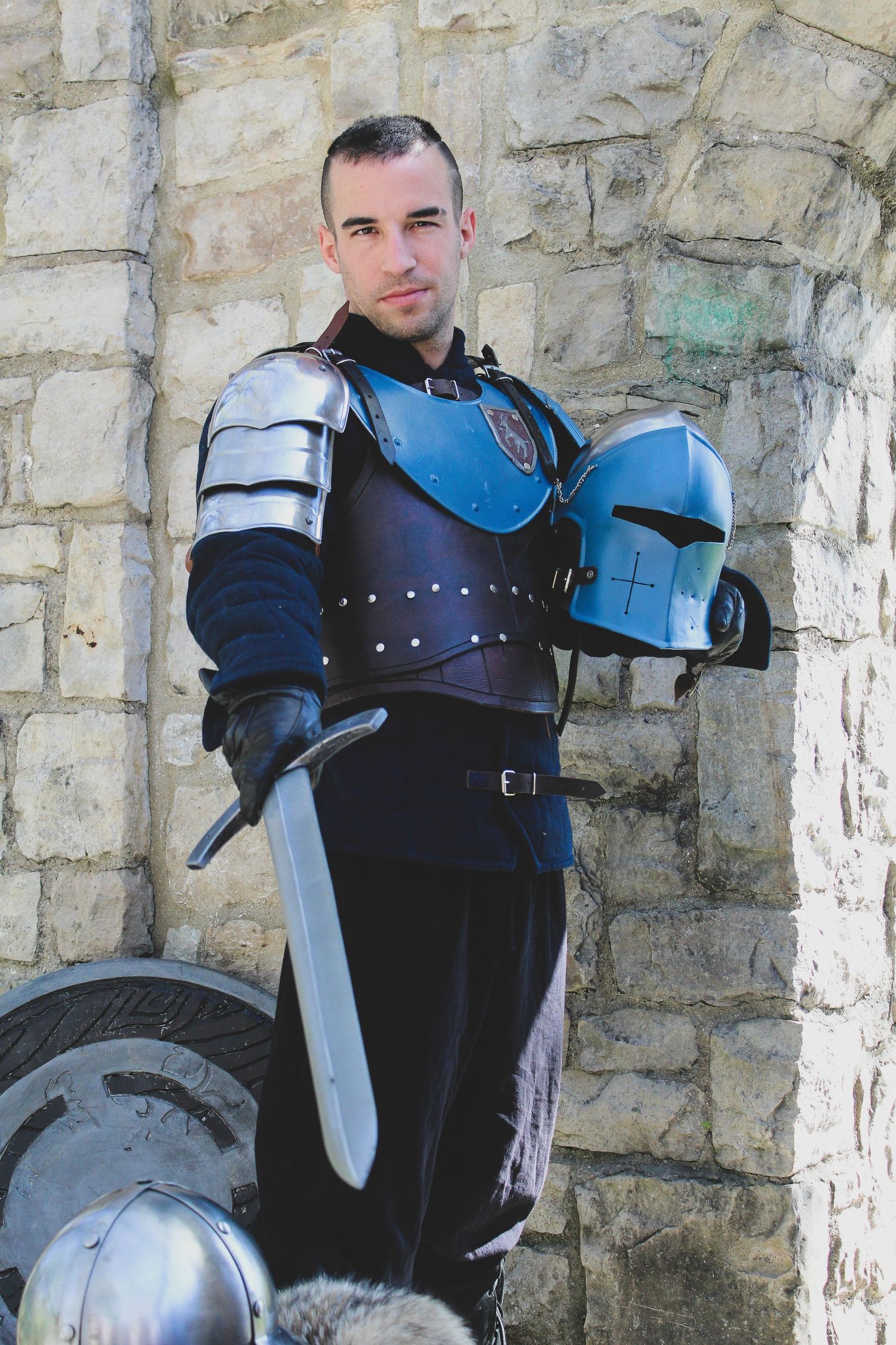 Medieval knight in blue and silver armor holds helmet and sword while standing against stone castle wall.