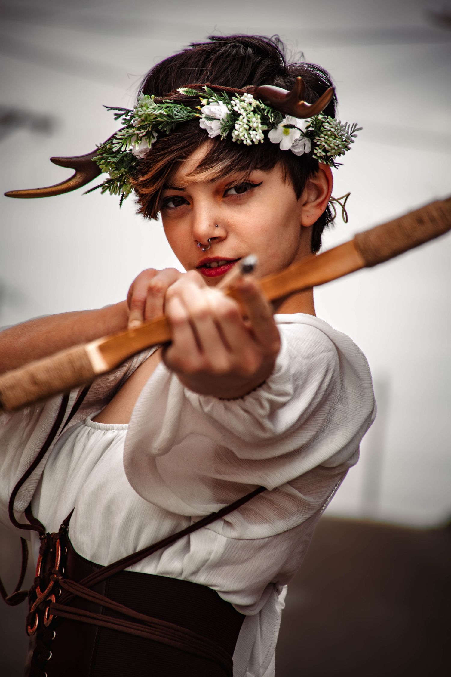 A person in a white shirt and floral crown aims a traditional wooden bow and arrow in a series of action shots.