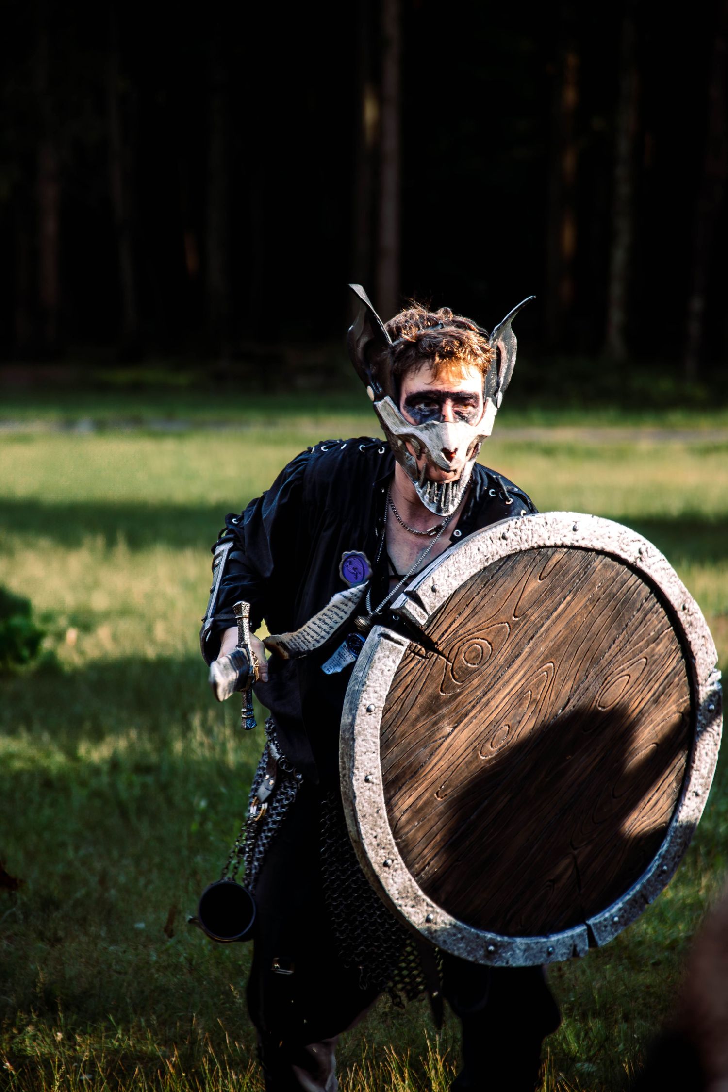 Person in medieval warrior costume with horned mask and wooden shield at outdoor LARP event.
