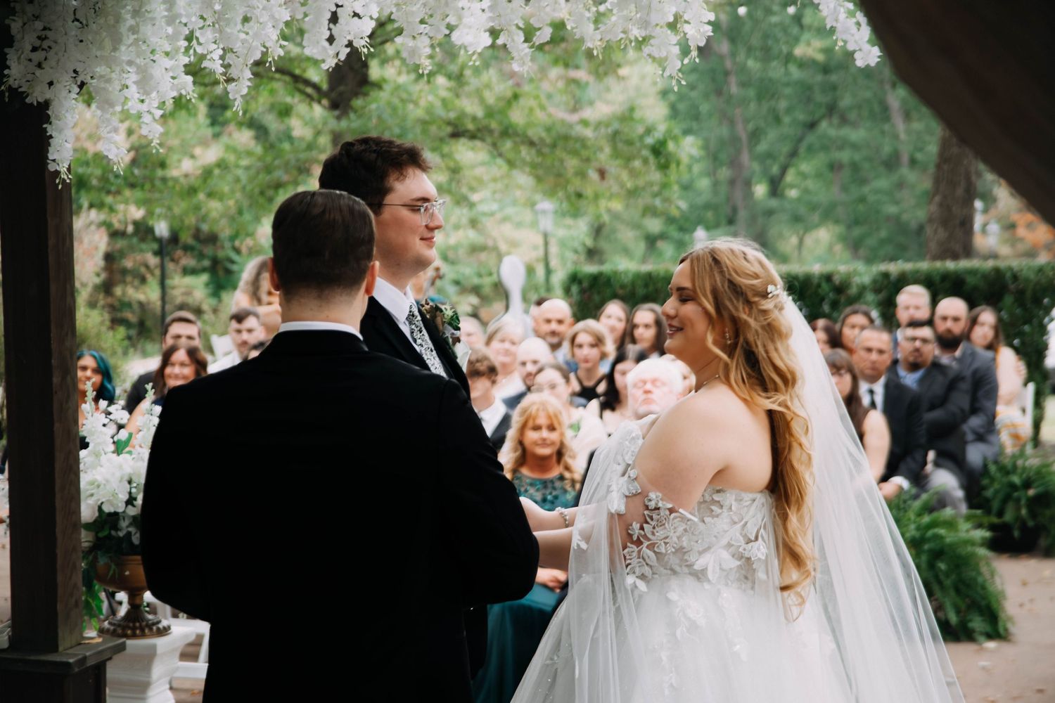Wedding celebration outdoors with guests seated under string lights and greenery.