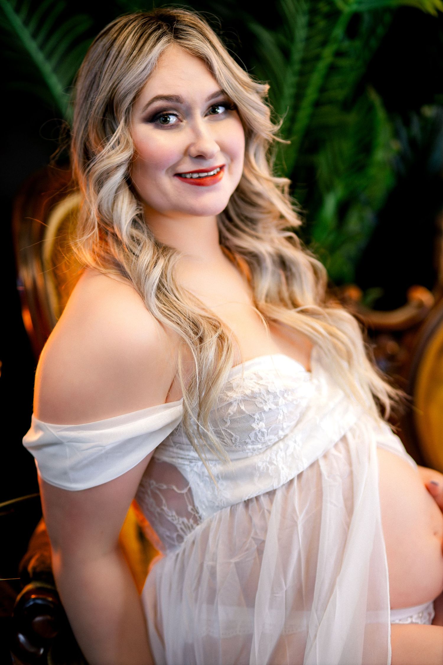 Pregnant woman in white lace off-shoulder gown with blonde wavy hair and red lips, posing elegantly in ornate chair.