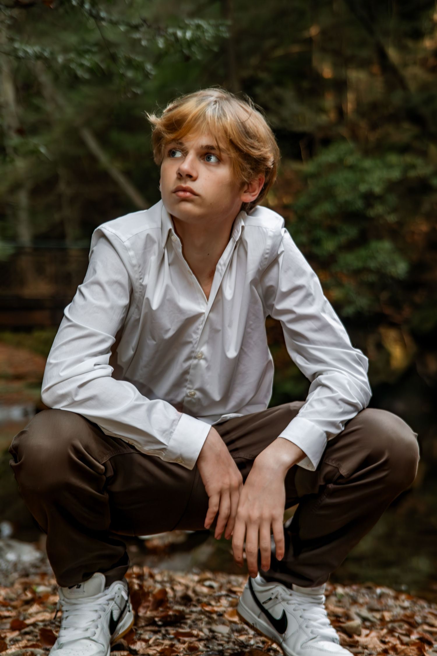Person in white button-down shirt and brown pants crouches among autumn leaves in a forest setting.